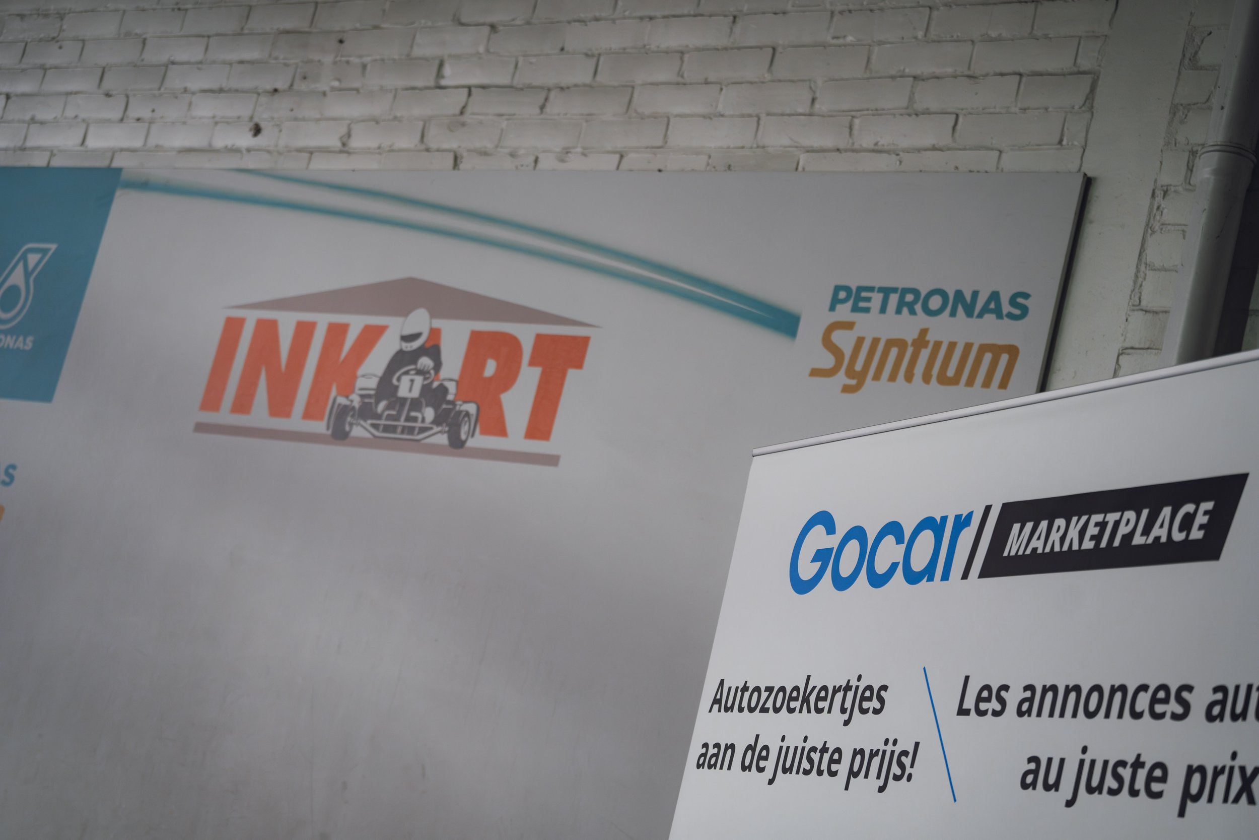 A partial view of a promotional banner on a white brick wall, featuring logos and text for automotive companies including Petronas Syntium and Gocar Marketplace, with an illustration of a person racing in a go-kart.