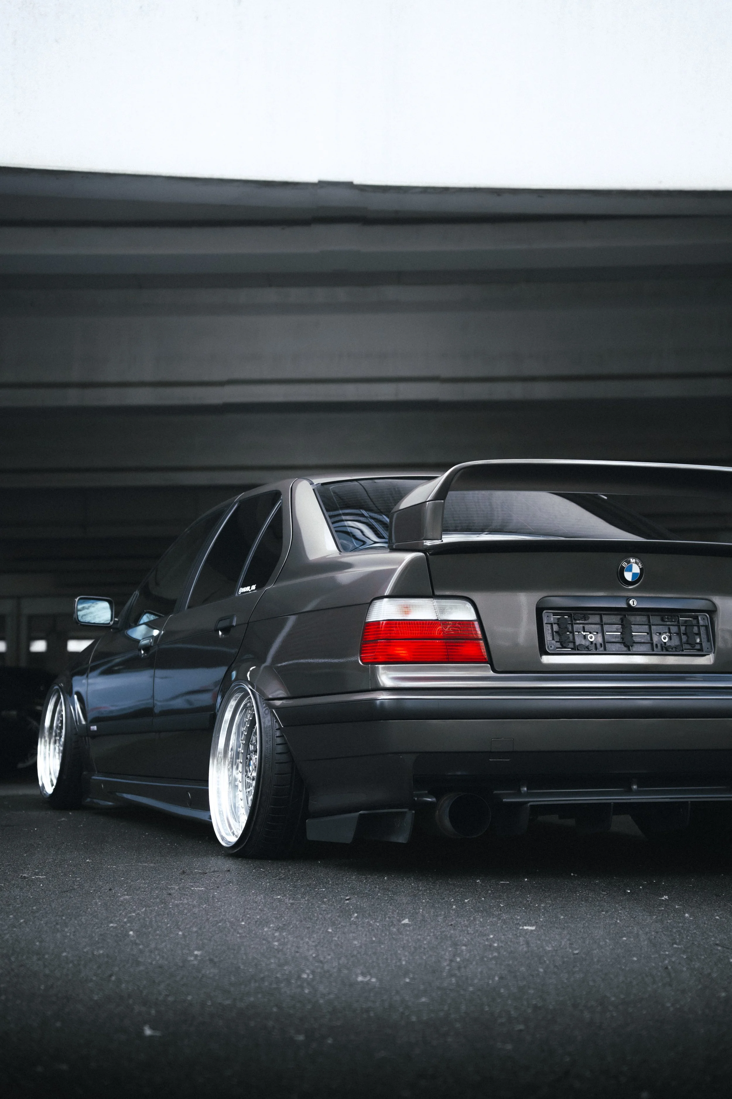 A gray BMW car with aftermarket wheels is parked in an indoor parking garage under a dark ceiling.