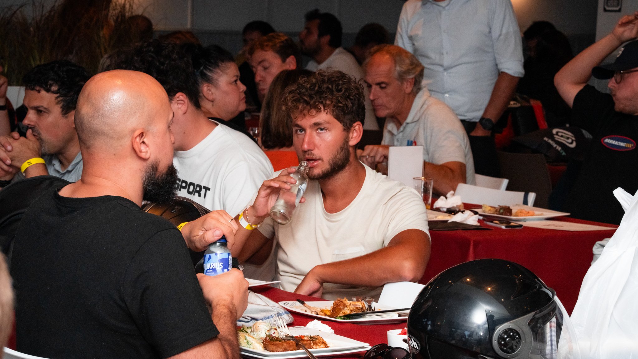 Young man with curly hair and beard drinking from a glass bottle at a crowded indoor event. He is sitting at a table with food, and a black helmet is in the foreground. Other people are seated at tables in the background.