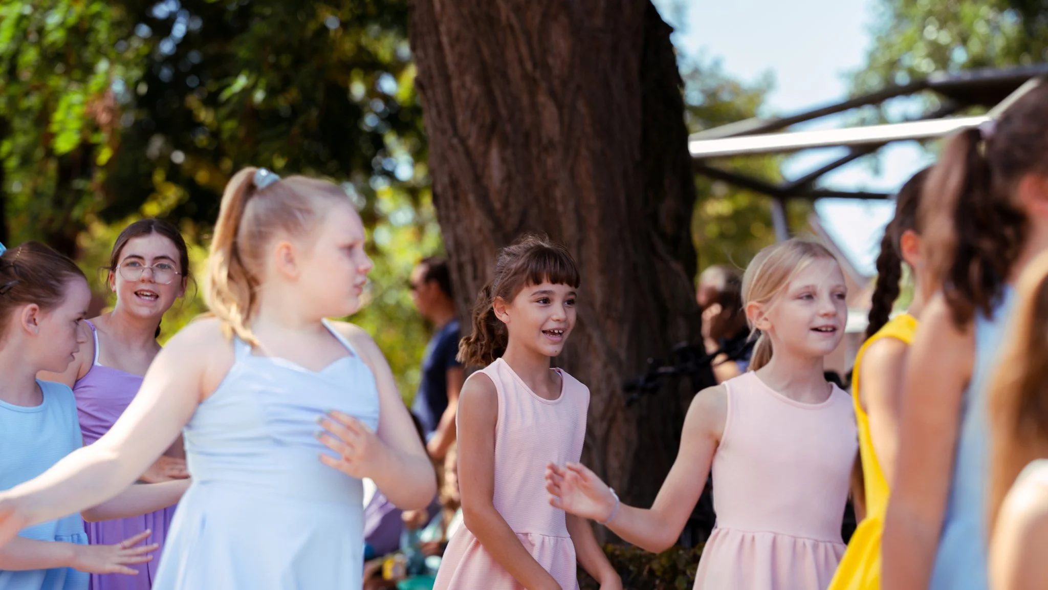 A group of young girls in pastel dresses standing and talking outside near a large tree on a sunny day.
