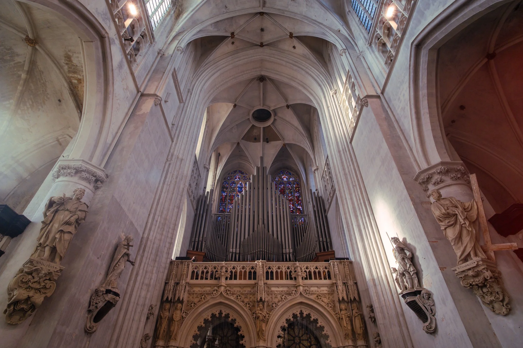 Interior view of a Gothic cathedral with high vaulted ceilings, stained glass windows, pipe organ, and statues along the walls.