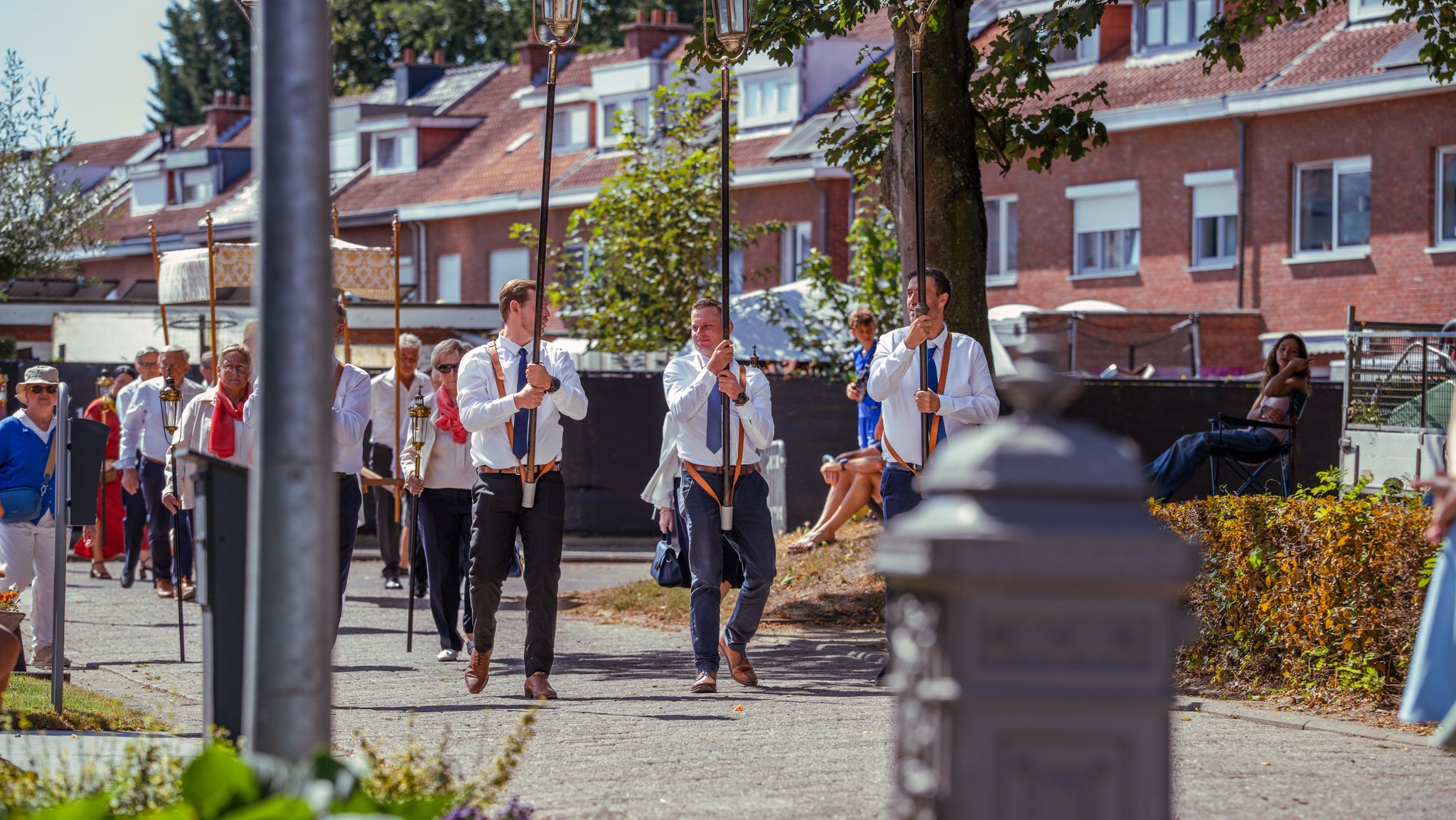 Group of people participating in a parade or procession outdoors, walking along a sidewalk with houses in the background, some holding lanterns and sticks, with spectators seated on the right side.