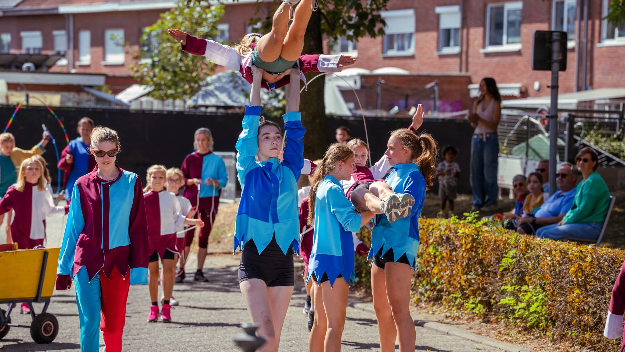A group of children performing a cheerleading stunt outdoors during daytime. Two girls are holding up a girl who is doing a handstand with her legs spread apart, while other children and adults watch.