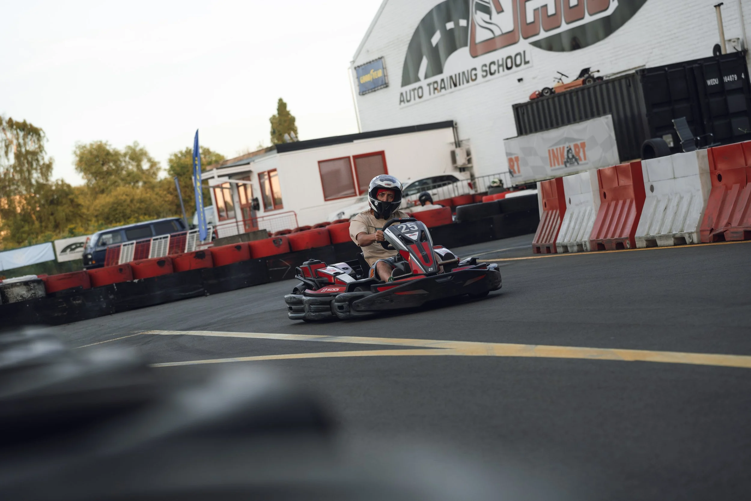 Person wearing a helmet driving a go-kart on a racetrack at an auto training school, with barriers and a large white building in the background.