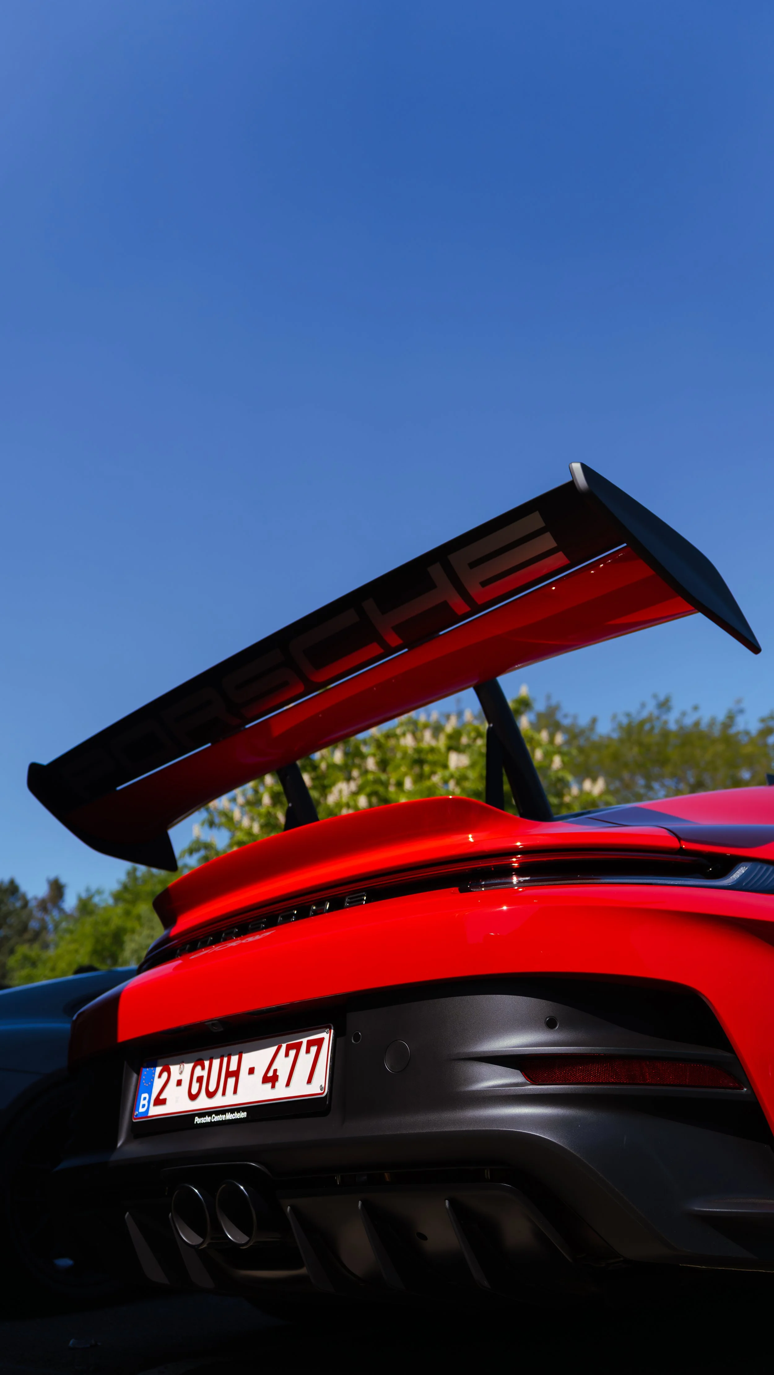 Close-up of the rear of a red Porsche 911 sports car, showing the large rear wing, taillights, dual exhaust pipes, and license plate reading '2-GUH-477', against a clear blue sky and green trees in the background.