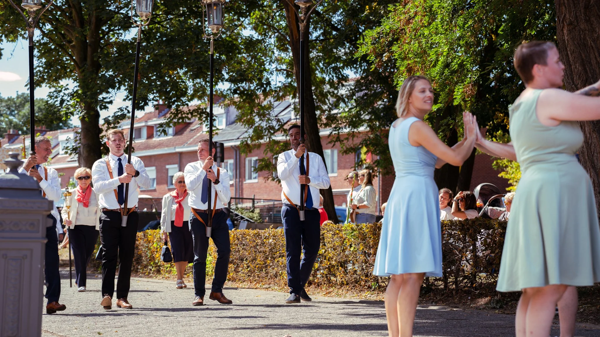 People participating in a memorial or commemorative event outdoors, with some exchanging high fives near a large tree, and a group of flag bearers in the background.