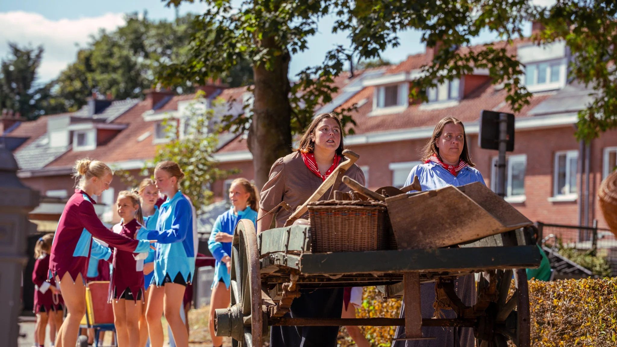 Two women dressed as musicians performing with a cart in front of young dancers in colorful costumes during an outdoor street performance.