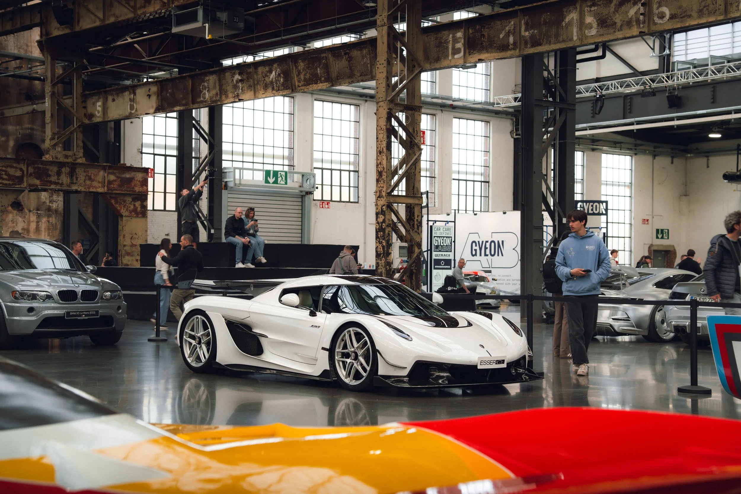 Several luxury cars, including a white sports car and a silver BMW, on display at an indoor car exhibition with people walking around and looking at the vehicles.