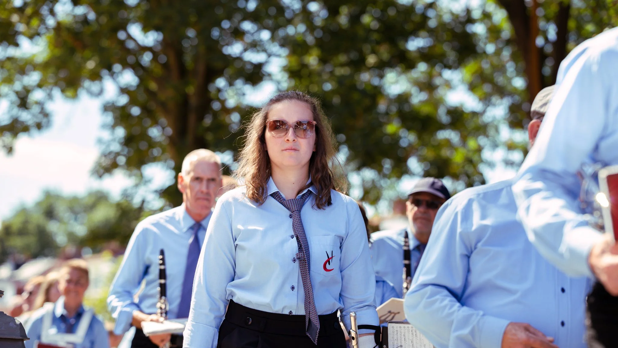 Women in light blue shirts participating in an outdoor musical event, holding sheet music, under a large leafy tree.