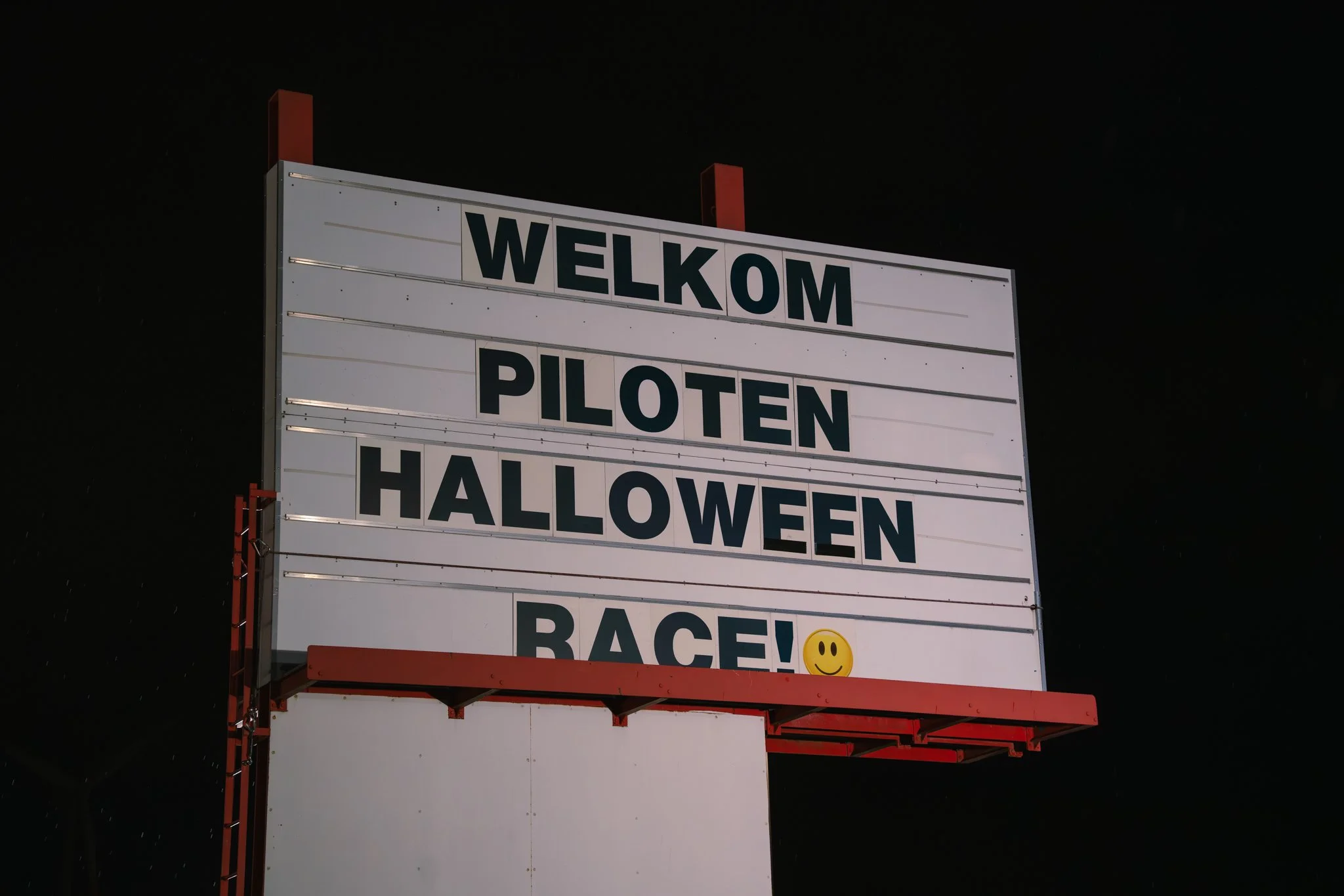 A large signboard at night that says "Welkom Piloten Halloween Race" with a smiley face emoji.
