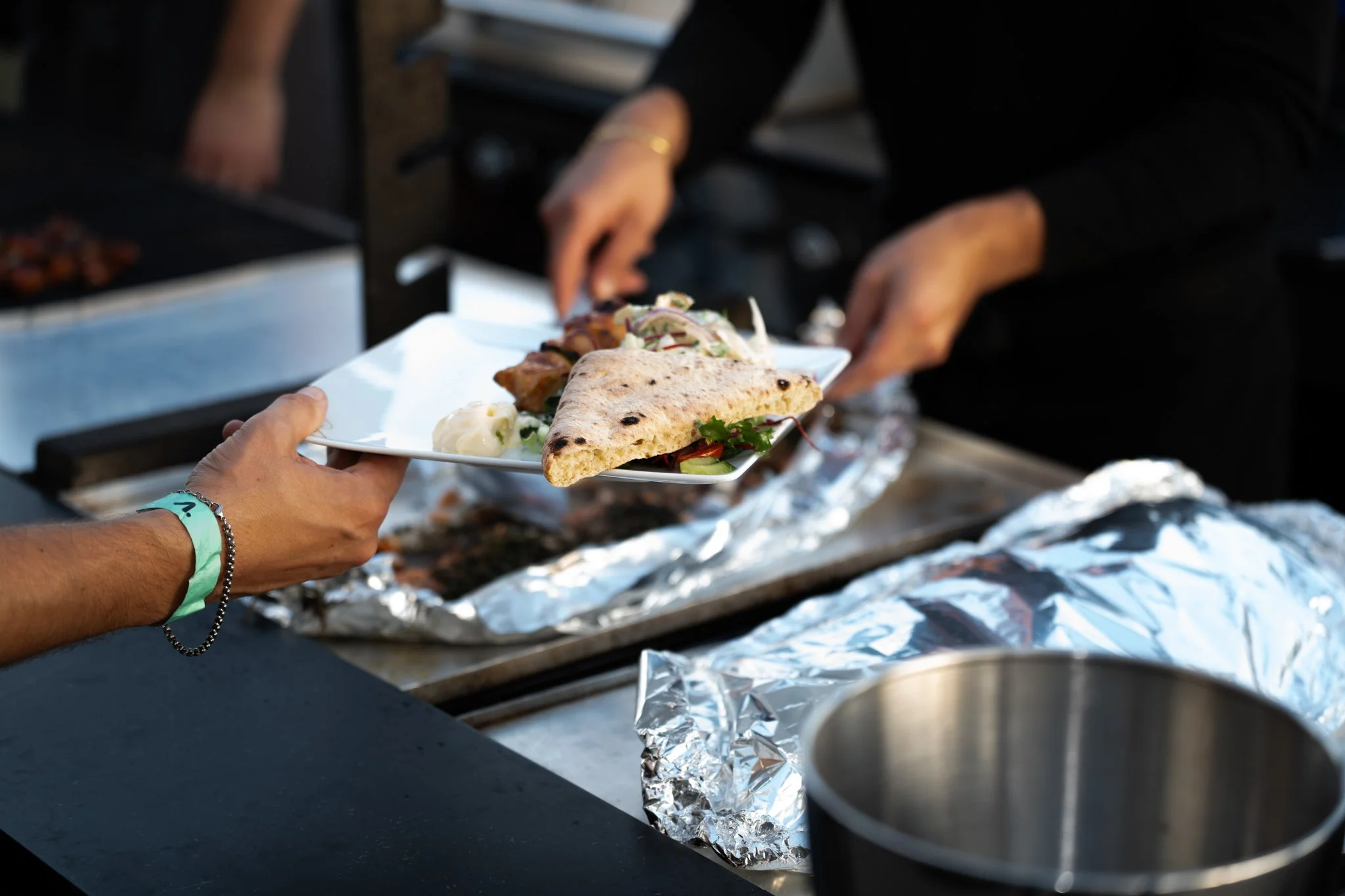 Person handing a plate of food, including naan bread and salad, to another person at a food stand with foil-covered grill in the background.