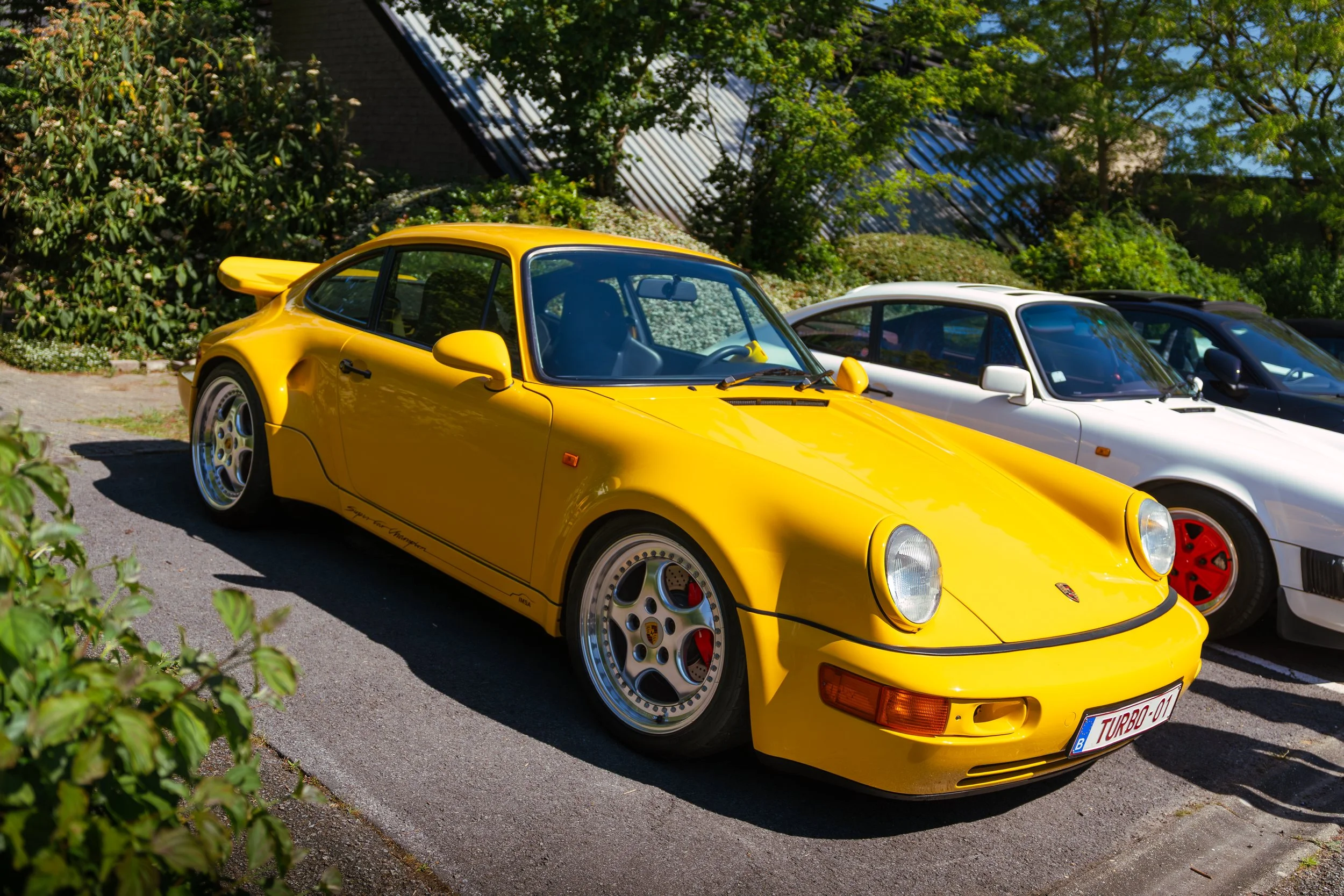 Yellow vintage Porsche 911 sports car parked outdoors, with a white car beside it, and green trees in the background.