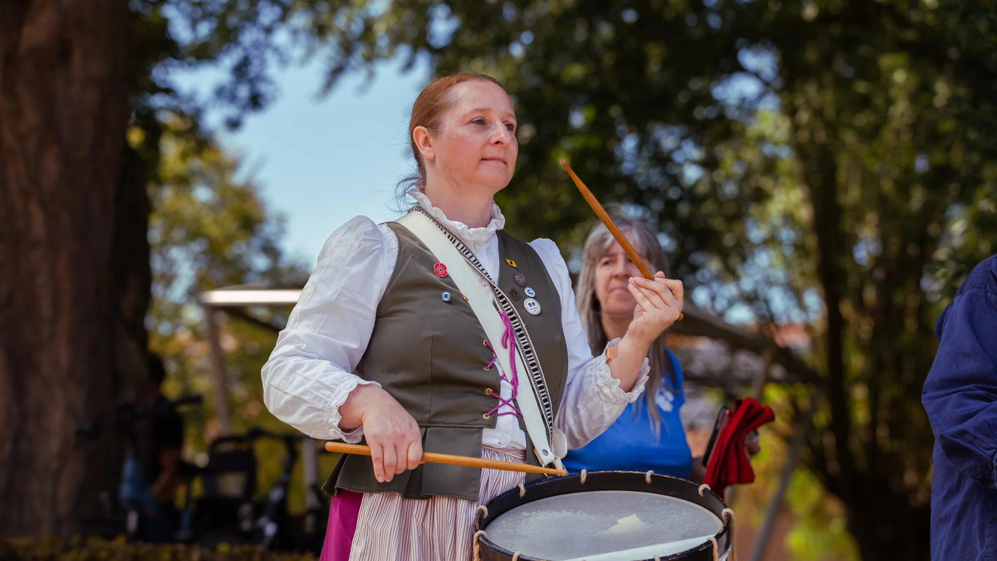 A woman dressed in traditional attire holding a drum and drumsticks, participating in an outdoor cultural event