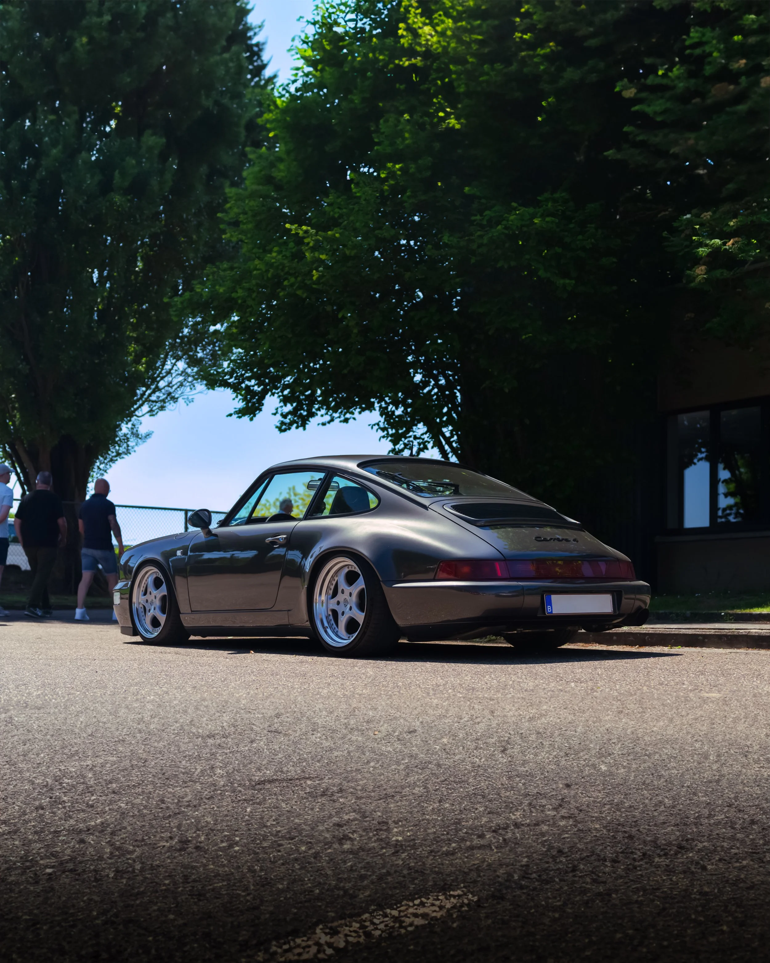 A classic black Porsche 911 parked on a street with large trees and a building in the background, with a few people walking nearby.