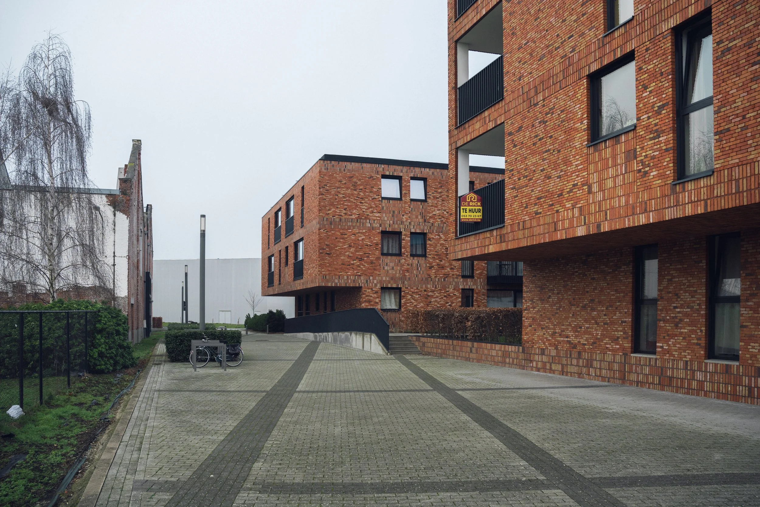 Several modern brick apartment buildings with black balconies and windows, a paved walkway, some bicycles, bushes, and a leafless tree on a cloudy day.