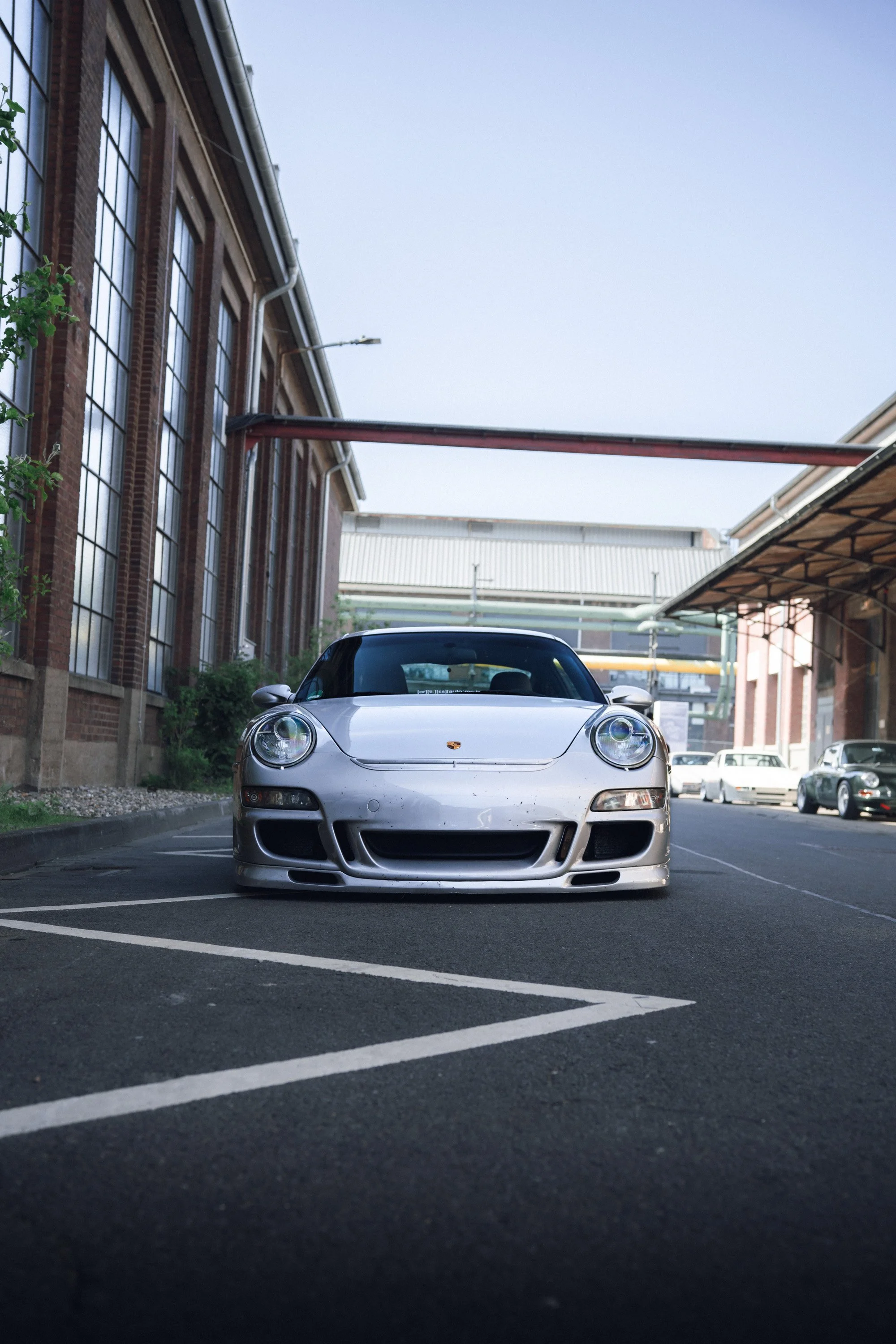 A silver Porsche sports car parked in an urban setting with industrial buildings and a cloudy sky in the background.
