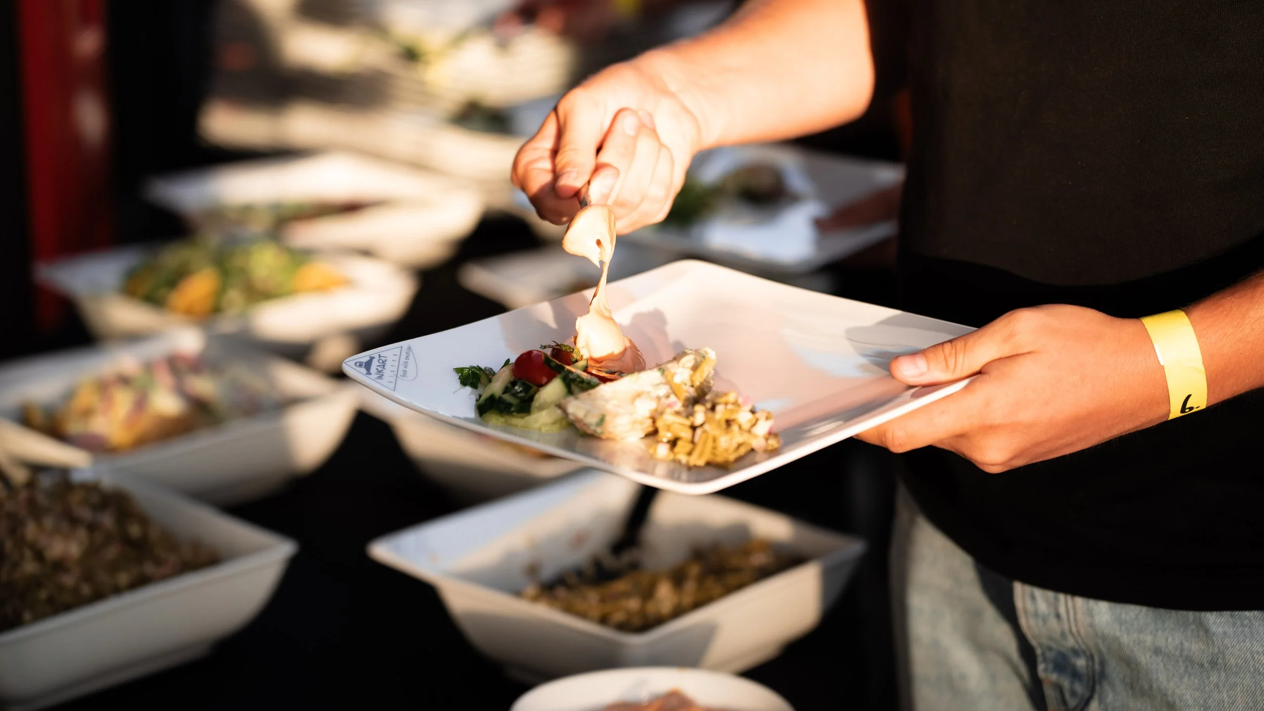 A person holding a white plate with a variety of salads and toppings, using a fork to add a piece of cheese, with other plates of food blurred in the background.