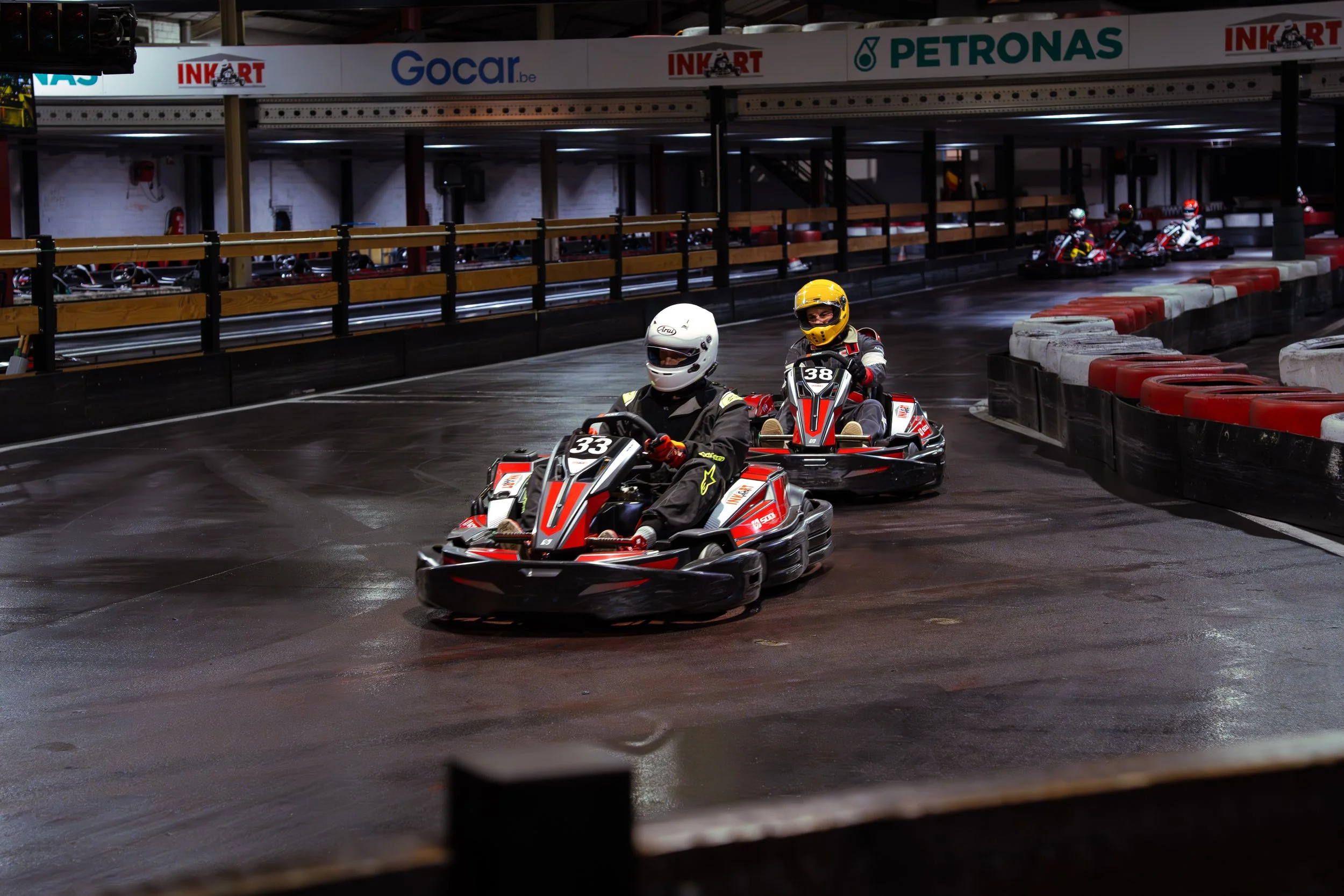 Two people in go-karts racing on an indoor track. Both are wearing helmets, with the person in front wearing a white helmet and the person behind in a yellow helmet. The track has a black surface with red and white barriers on the sides. In the backg