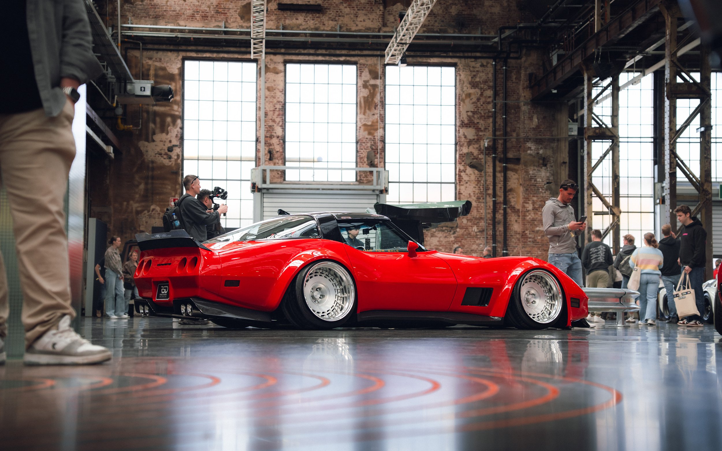 Red sports car displayed indoors at an exhibition, surrounded by people, with large industrial windows and exposed brick walls in the background.