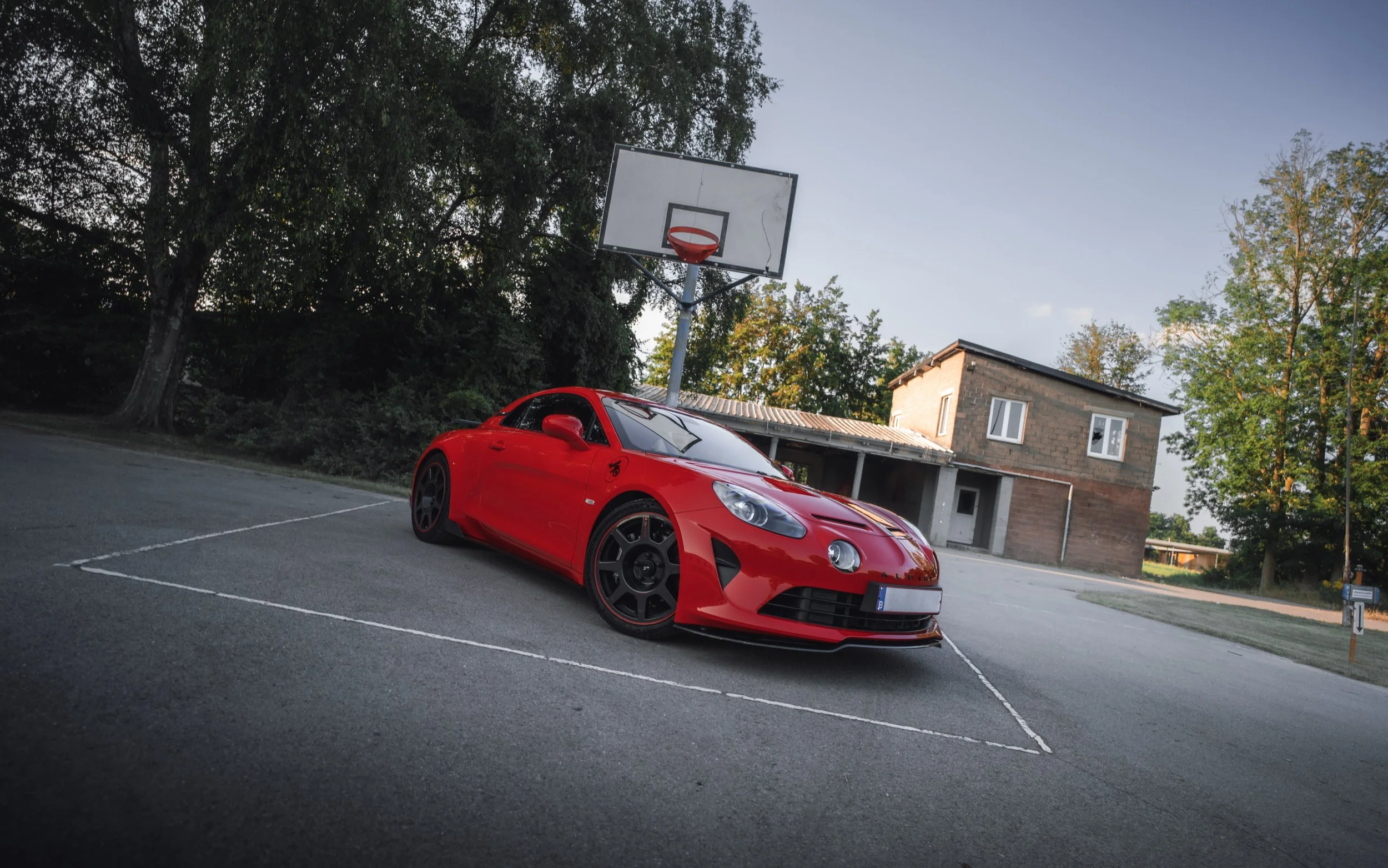 A red sports car parked on an empty outdoor basketball court with a basketball hoop and a residential building in the background, surrounded by trees, during daylight.