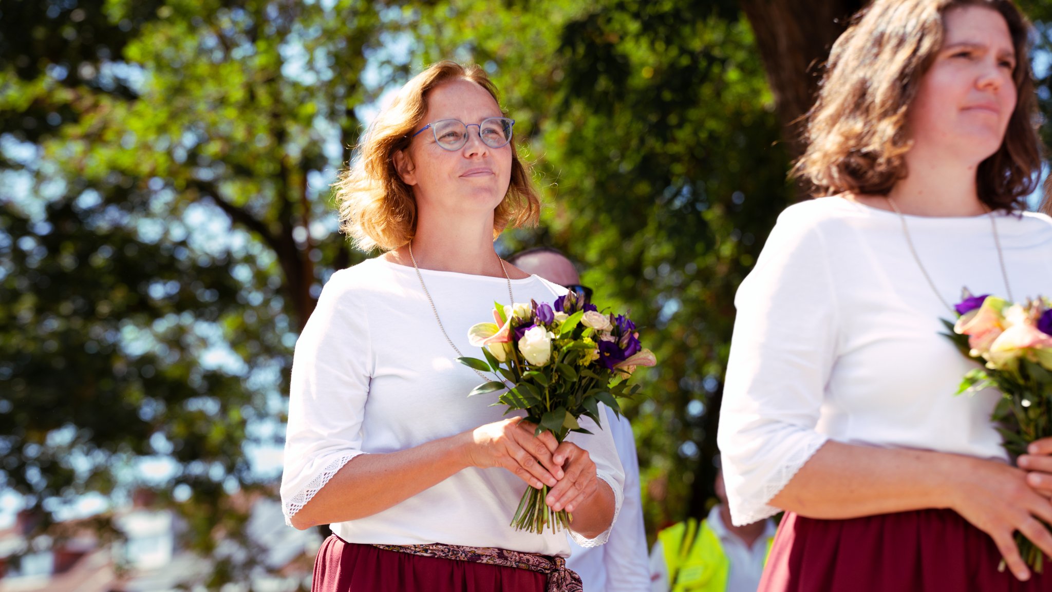 Women participating in an outdoor ceremony, holding bouquets of flowers, with trees in the background.