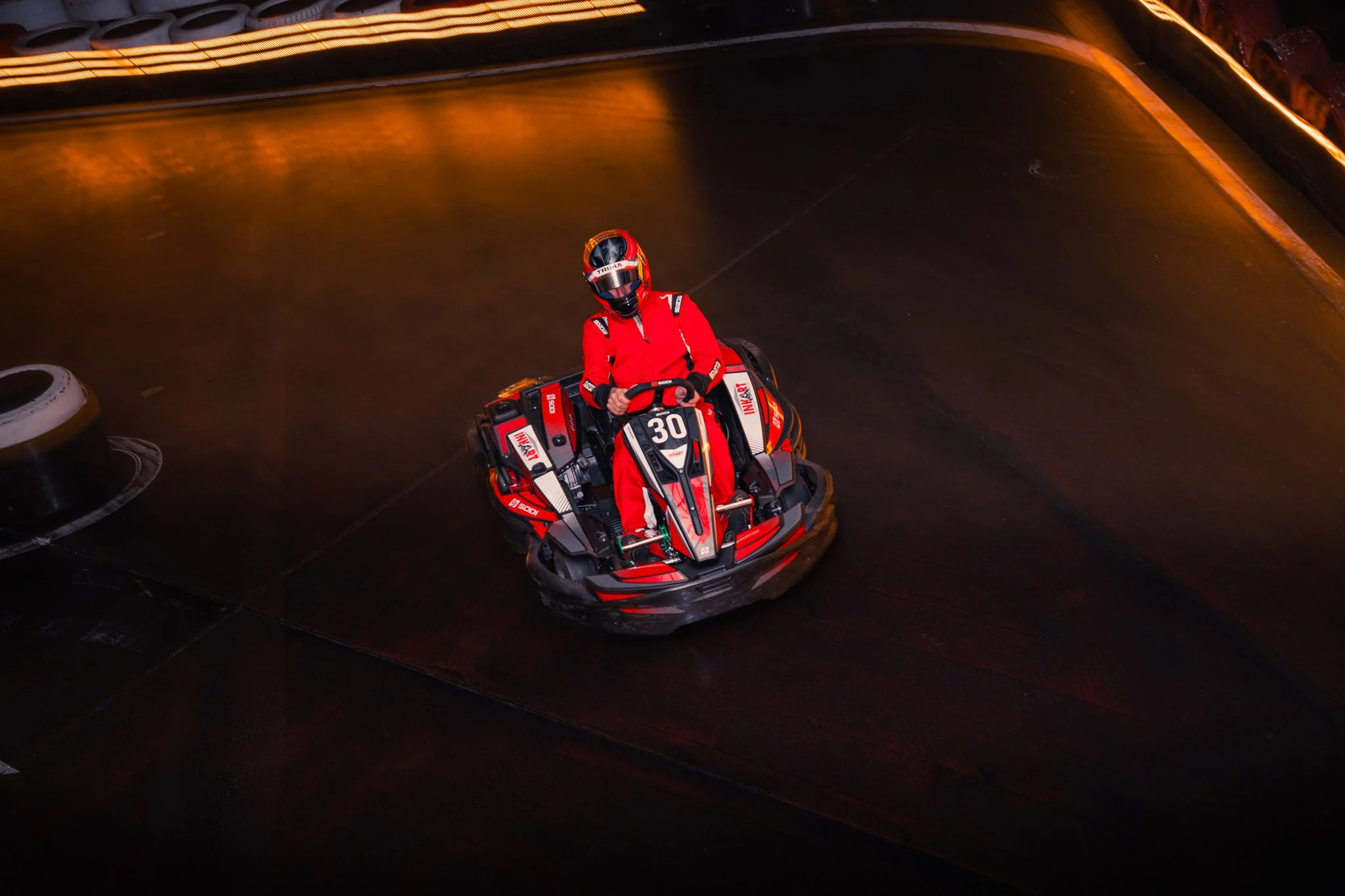 A person in a red racing suit and helmet riding a go-kart on an indoor track with dark flooring and orange lighting along the edges.