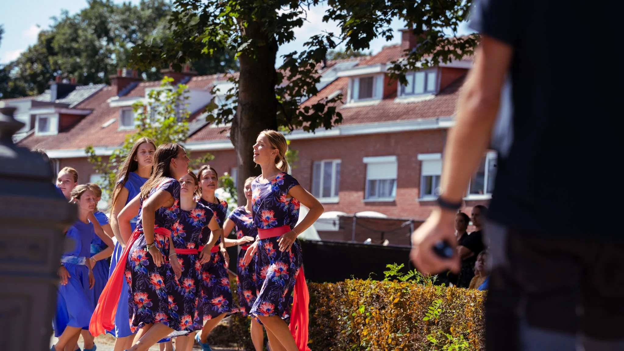 Group of women and girls dancing outdoors on a sunny day, with brick buildings and trees in the background.