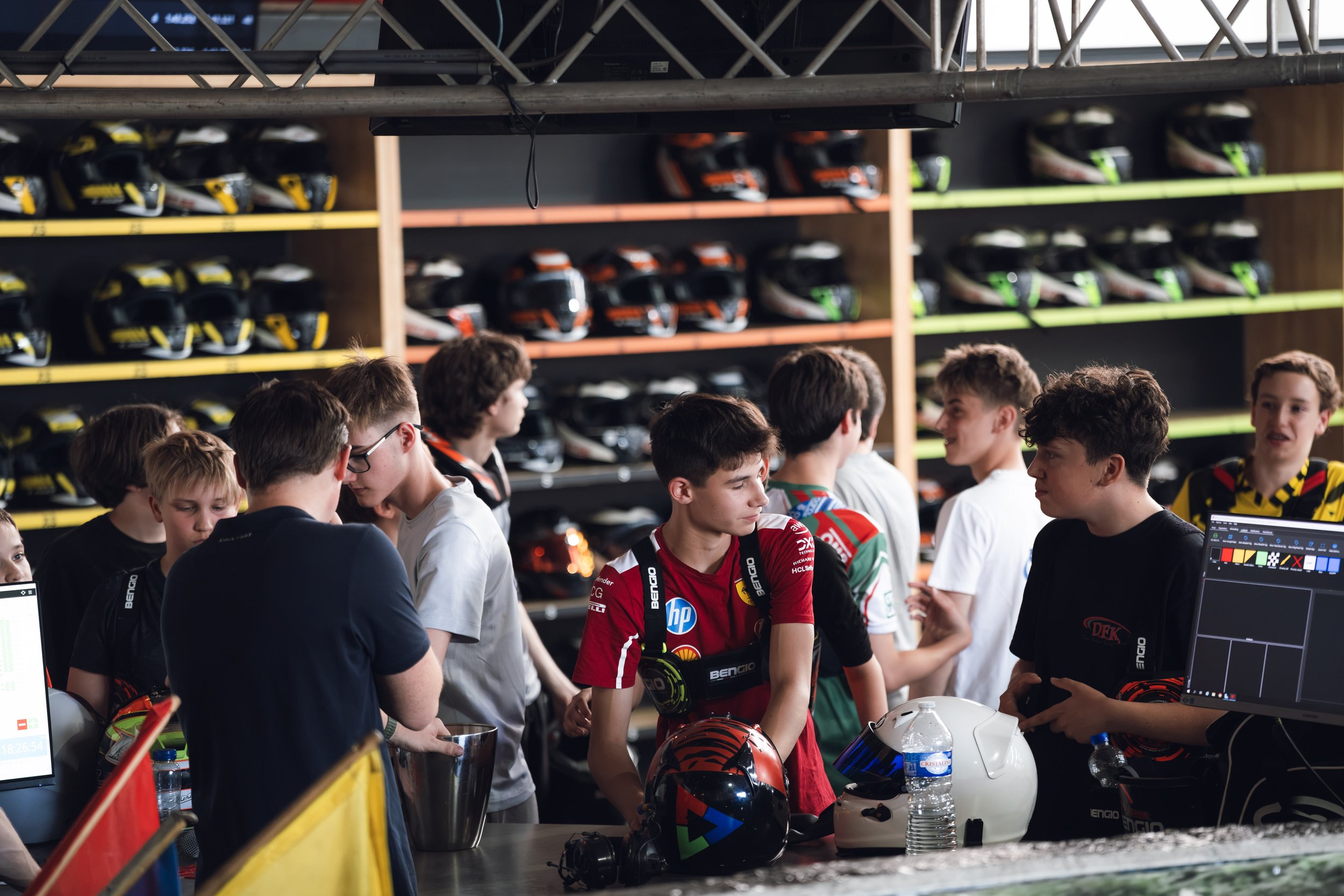 A group of young people, likely race car drivers, gathered around a table with helmets, water bottles, and computers, in a setting with shelves of helmets in the background.