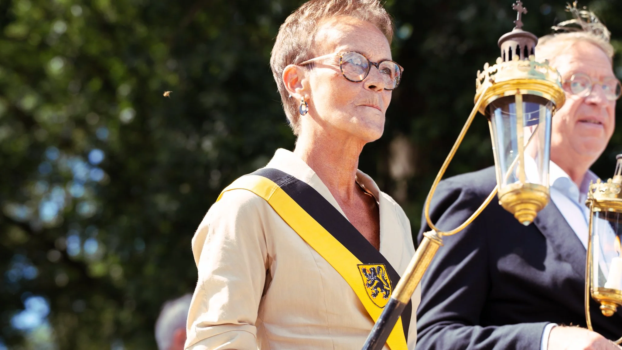 Two elderly individuals in formal attire participating in a public event, holding lanterns, with a woman wearing a sash with a crest, outdoors with trees in the background.