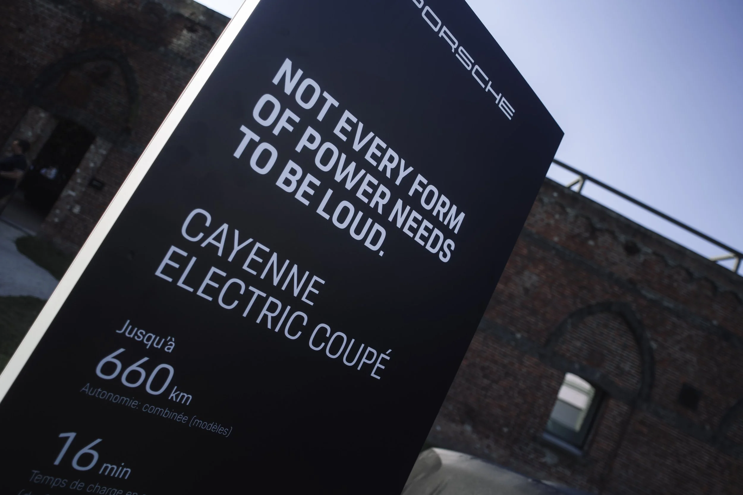 Signboard with information about electric vehicle charging, including distances and times, in front of a brick building.
