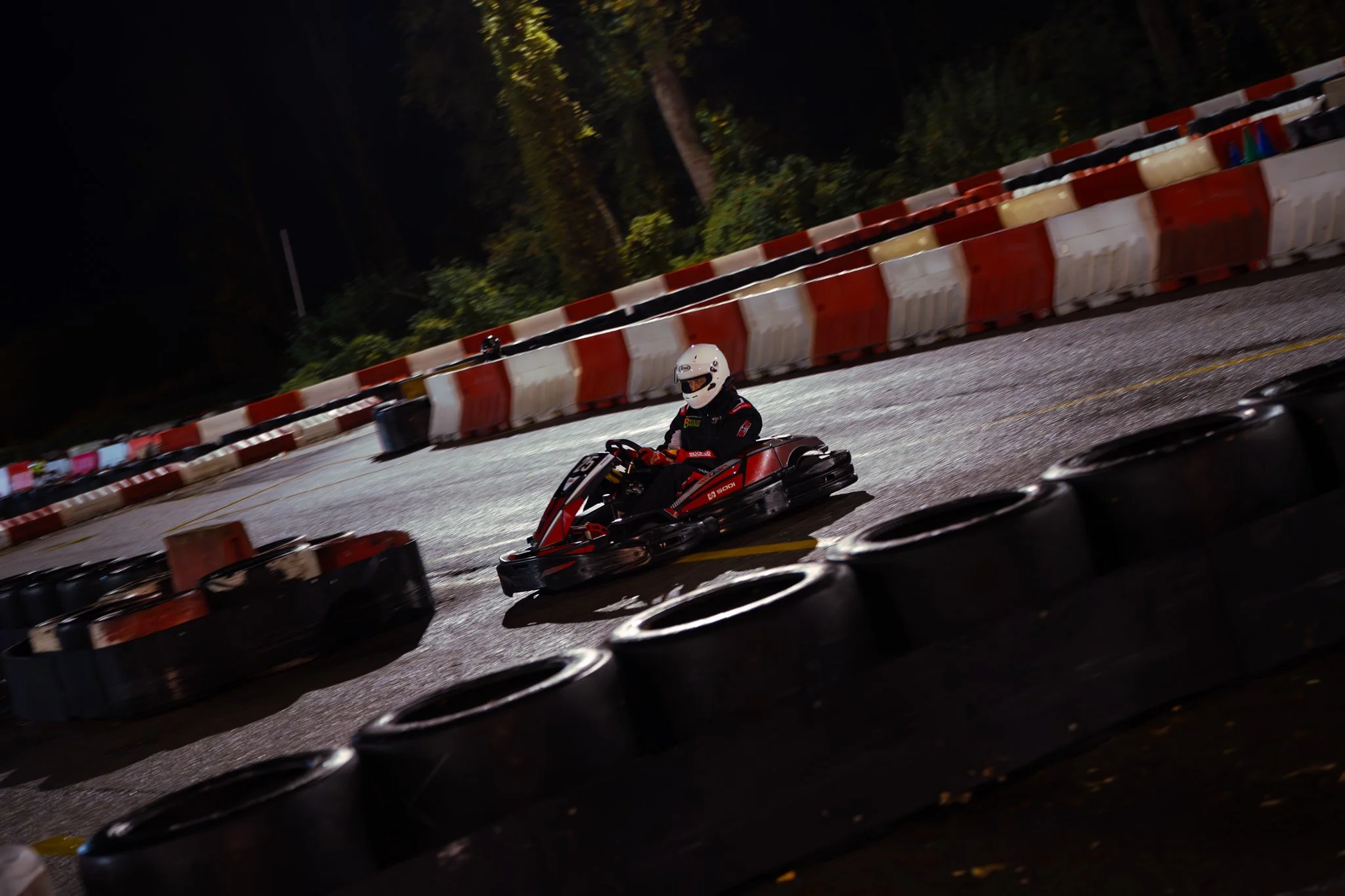 A person in racing gear and a helmet driving a go-kart on an outdoor track at night, surrounded by black and red tire barriers and orange and white safety barriers.