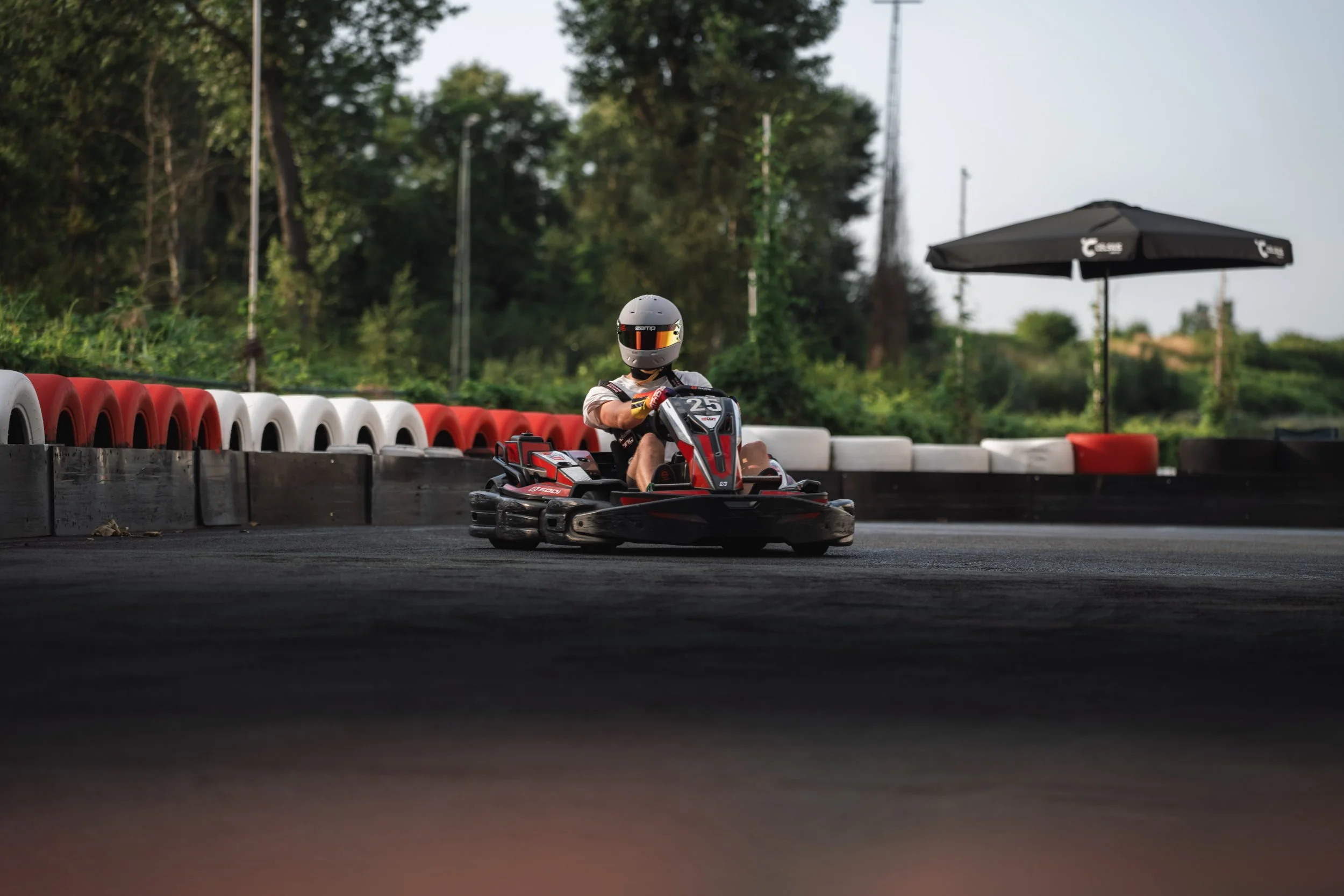 A person driving a go-kart on a track, wearing a helmet and gloves, with red and white tire barriers along the sides and trees in the background.