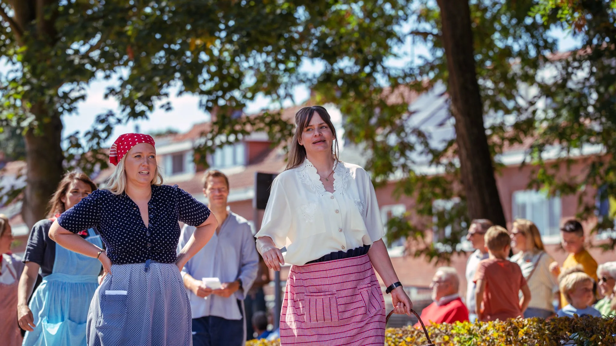 Women outdoors during a gathering or event, with trees and houses in the background, some people sitting and standing around.