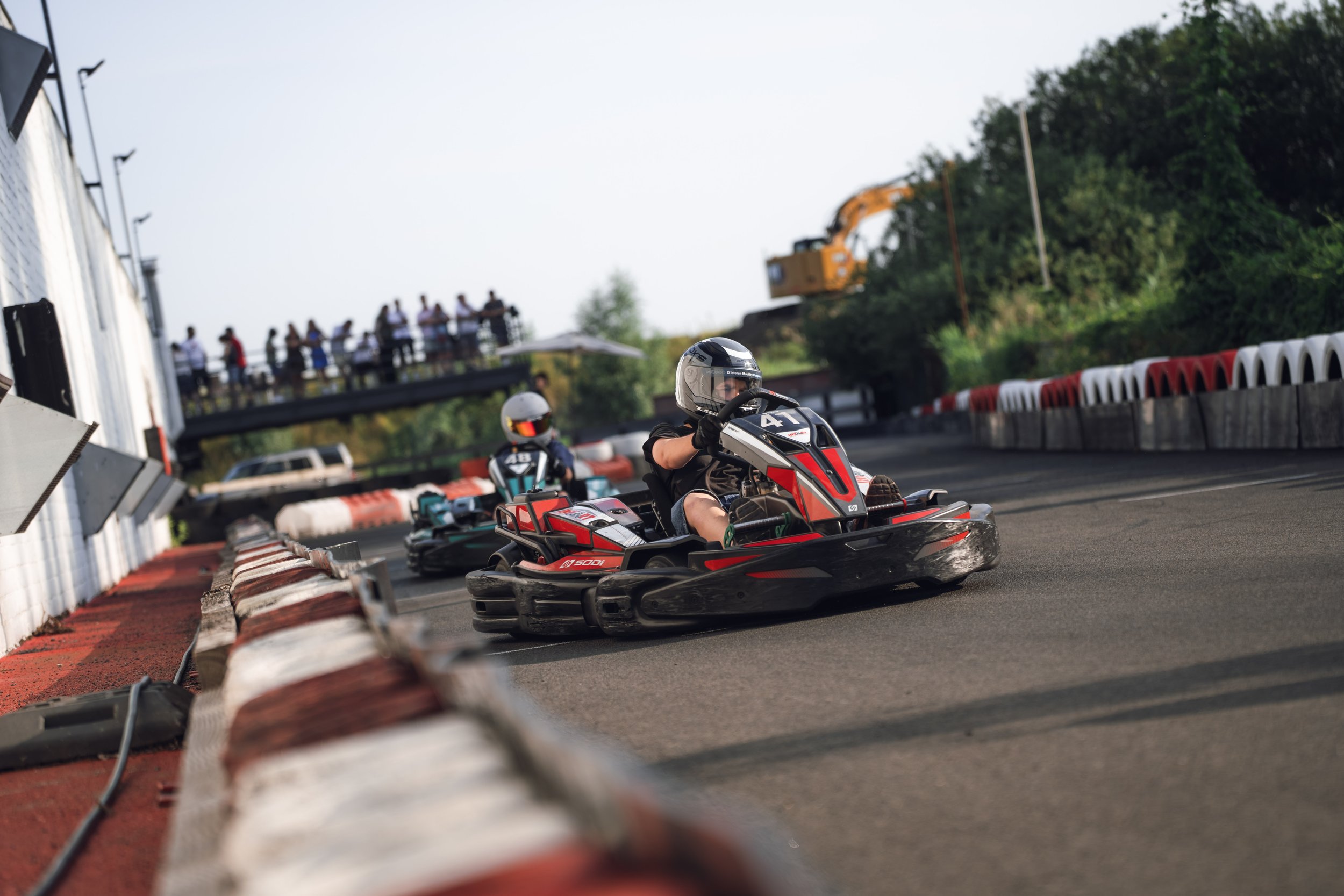 Two go-karts racing on an outdoor track with a red and white barrier, spectators watching from a bridge above, and a construction vehicle in the background surrounded by trees.