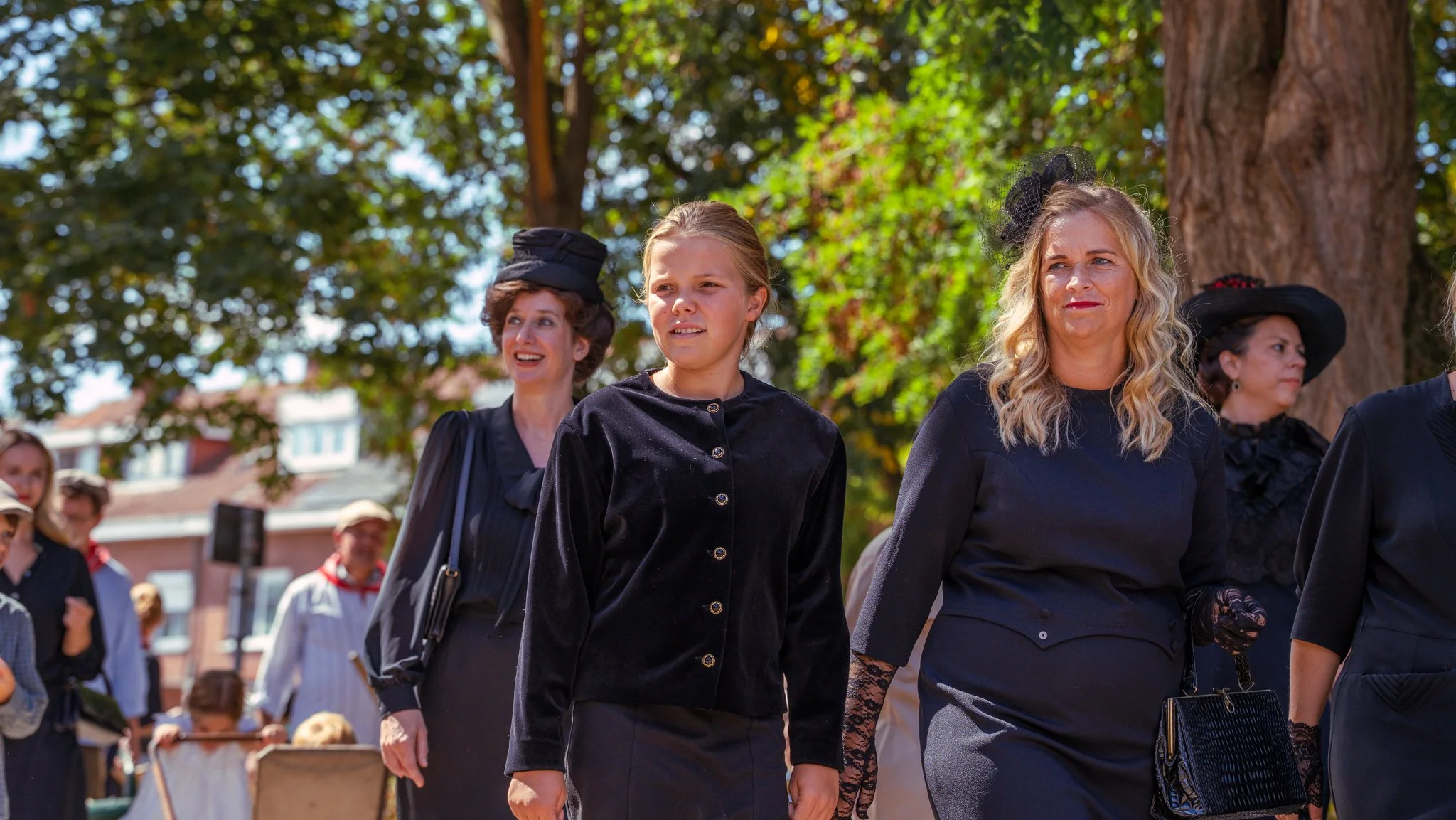 People dressed in vintage black attire walking outdoors in a park with large trees and buildings in the background.