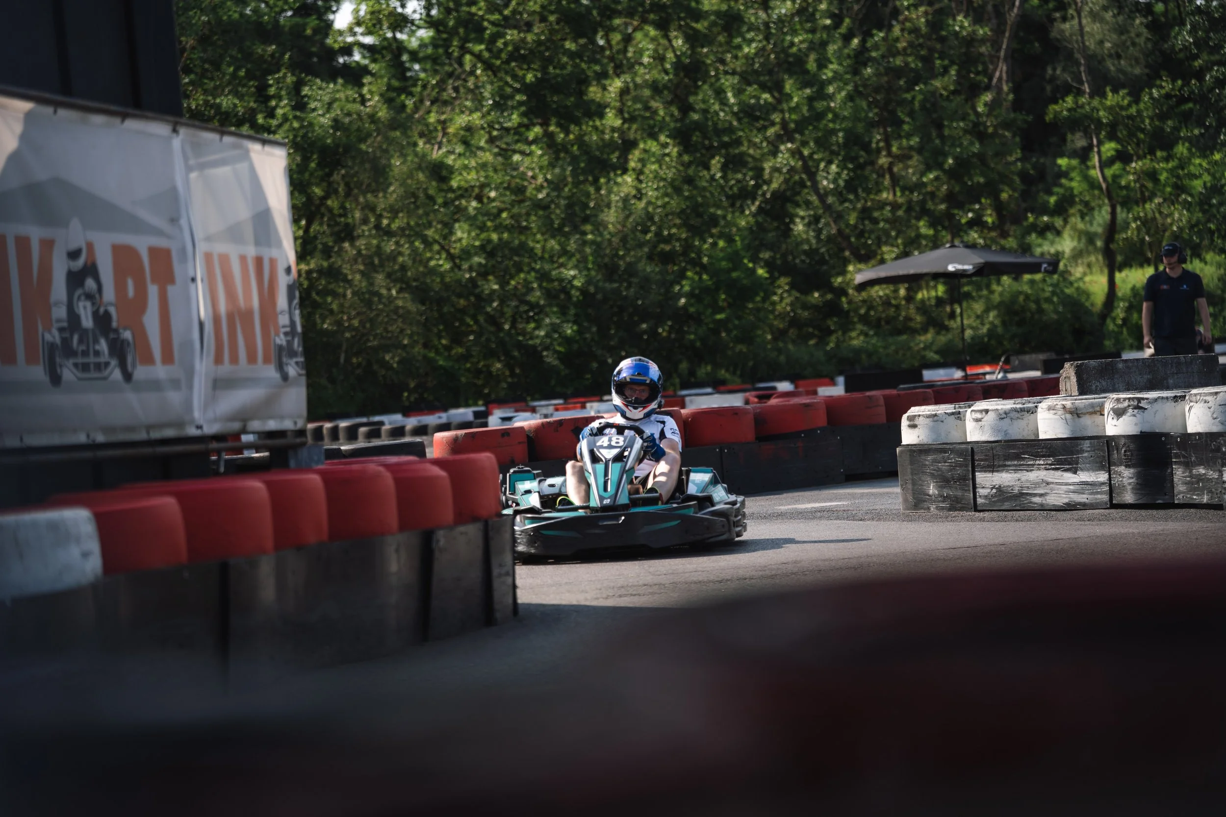 A person wearing a blue helmet driving a go-kart on an outdoor track lined with red and white barriers, with another track official standing nearby under a black umbrella, surrounded by green trees.