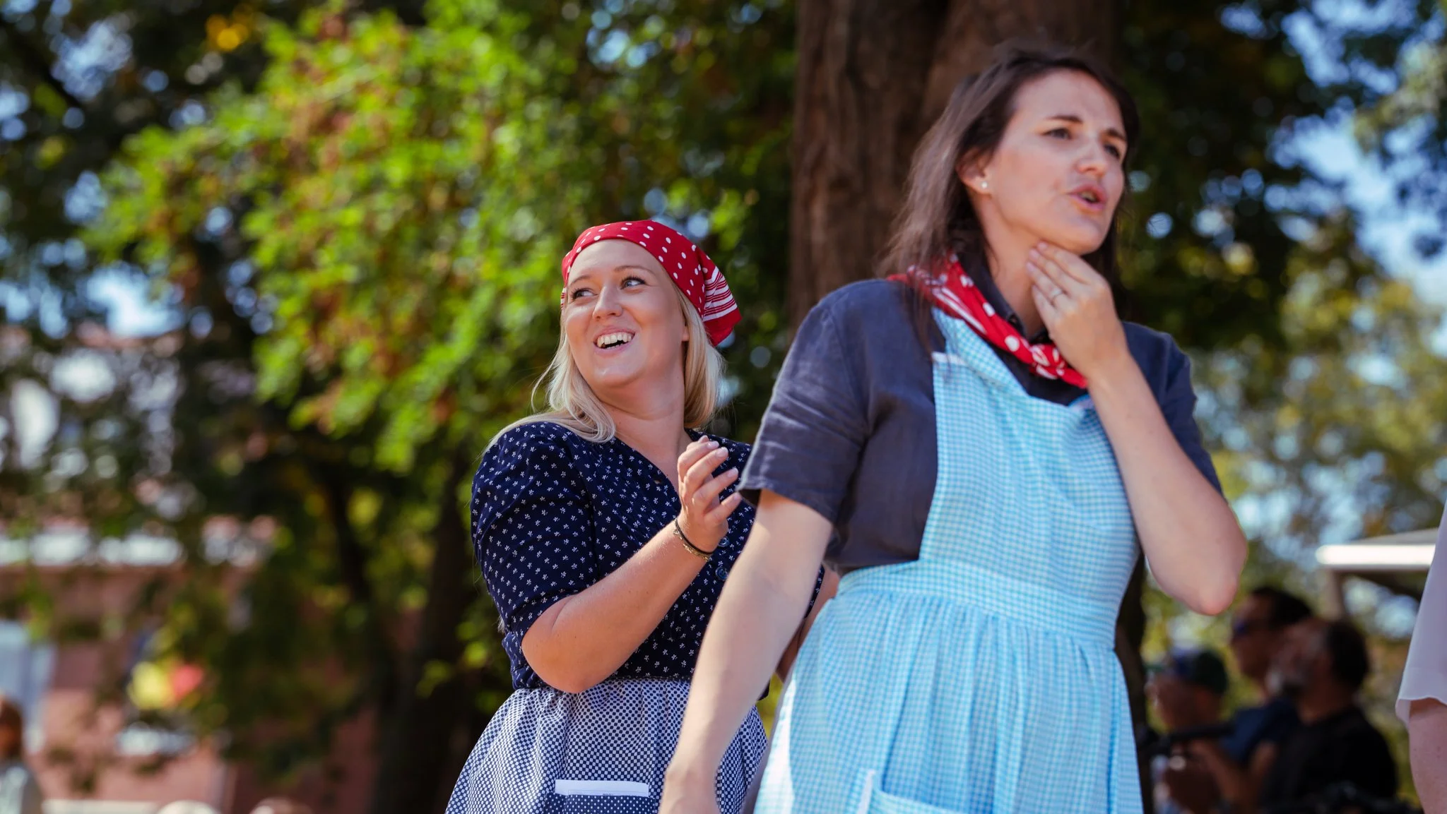 Two women in vintage aprons and bandanas standing outdoors near a large tree, smiling and chatting, with other people in the background.