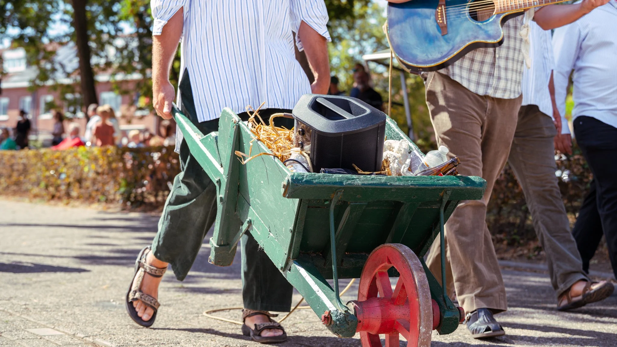 People walking outdoors pushing a green wheelbarrow filled with trash and a black portable speaker, with a guitarist walking beside them holding a guitar, and other people in the background.