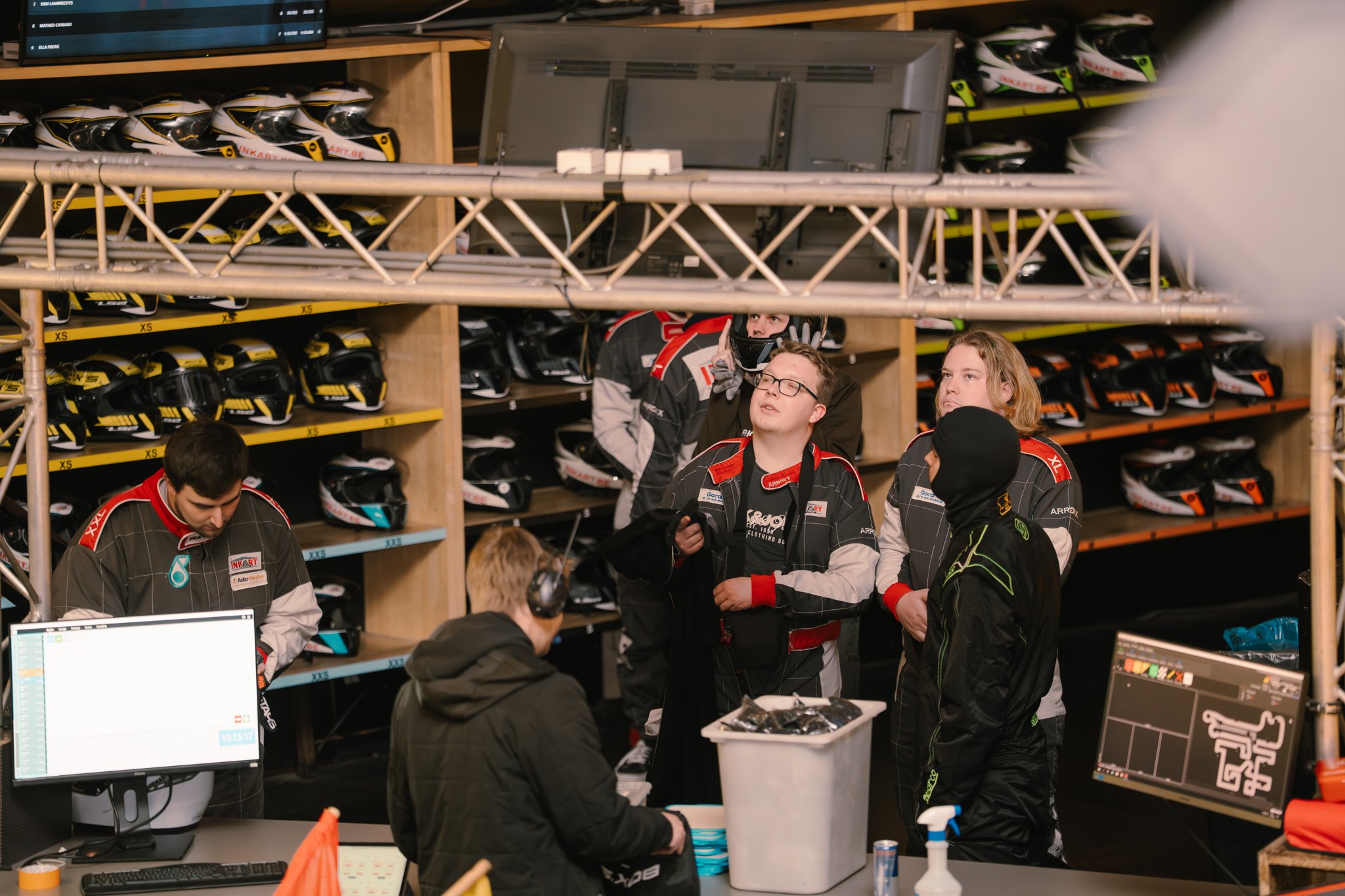 Race car team members in a garage, surrounded by helmets on shelves, discussing strategies. One team member is using a computer, and others are listening, all dressed in racing gear.