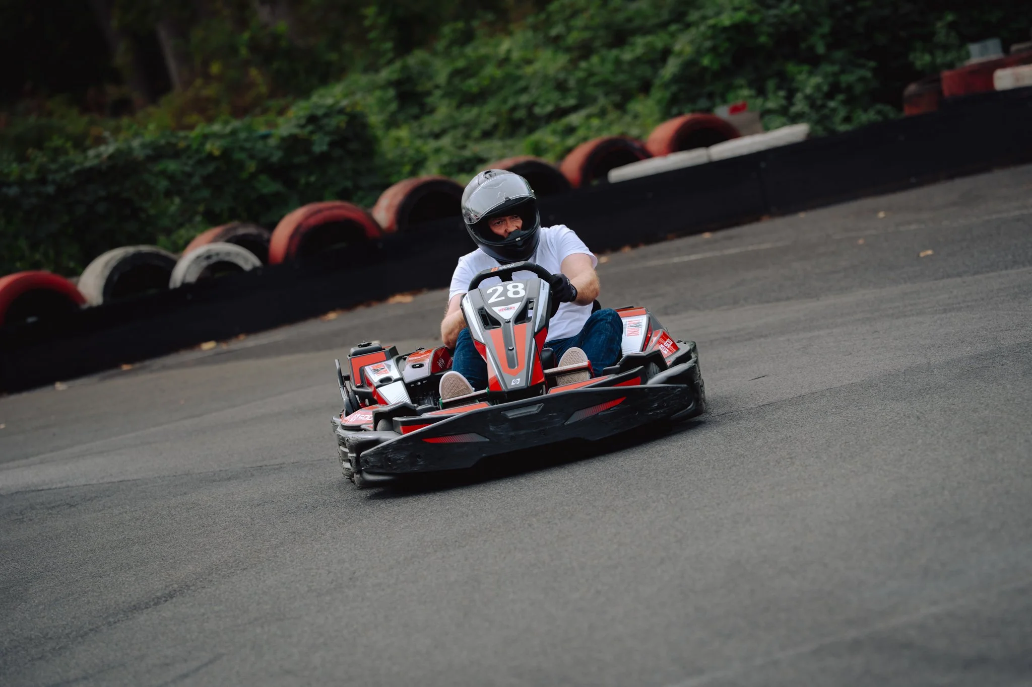 A person wearing a helmet and white t-shirt driving a go-kart on an outdoor race track with a black and red tire barrier in the background.