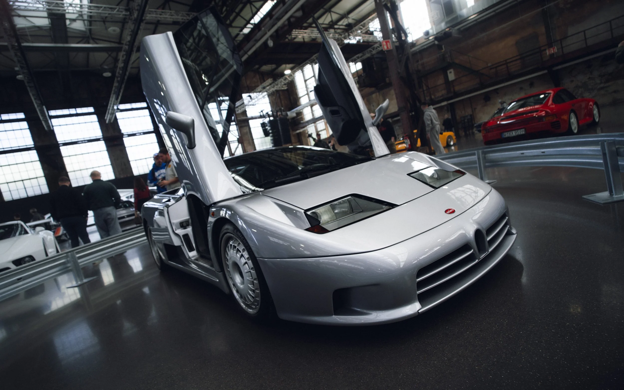 A silver sports car with its scissor doors open on display at an indoor car exhibition. In the background, other luxury cars and people are visible inside a large industrial-style building with high ceilings and large windows.