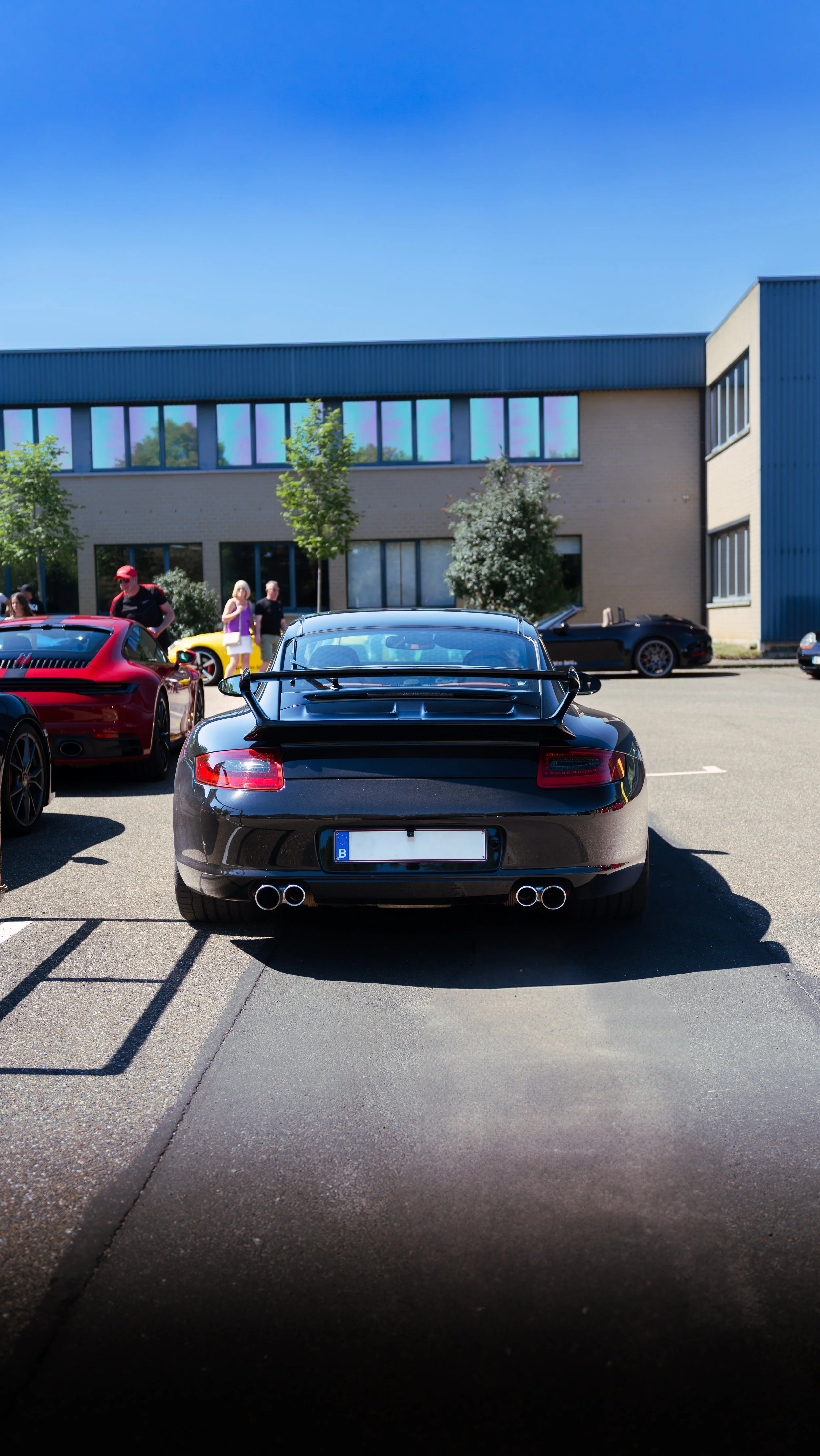 Black sports car with a rear spoiler and quad exhaust pipes parked in a lot at a car show, with people and other cars visible in the background under a clear blue sky.
