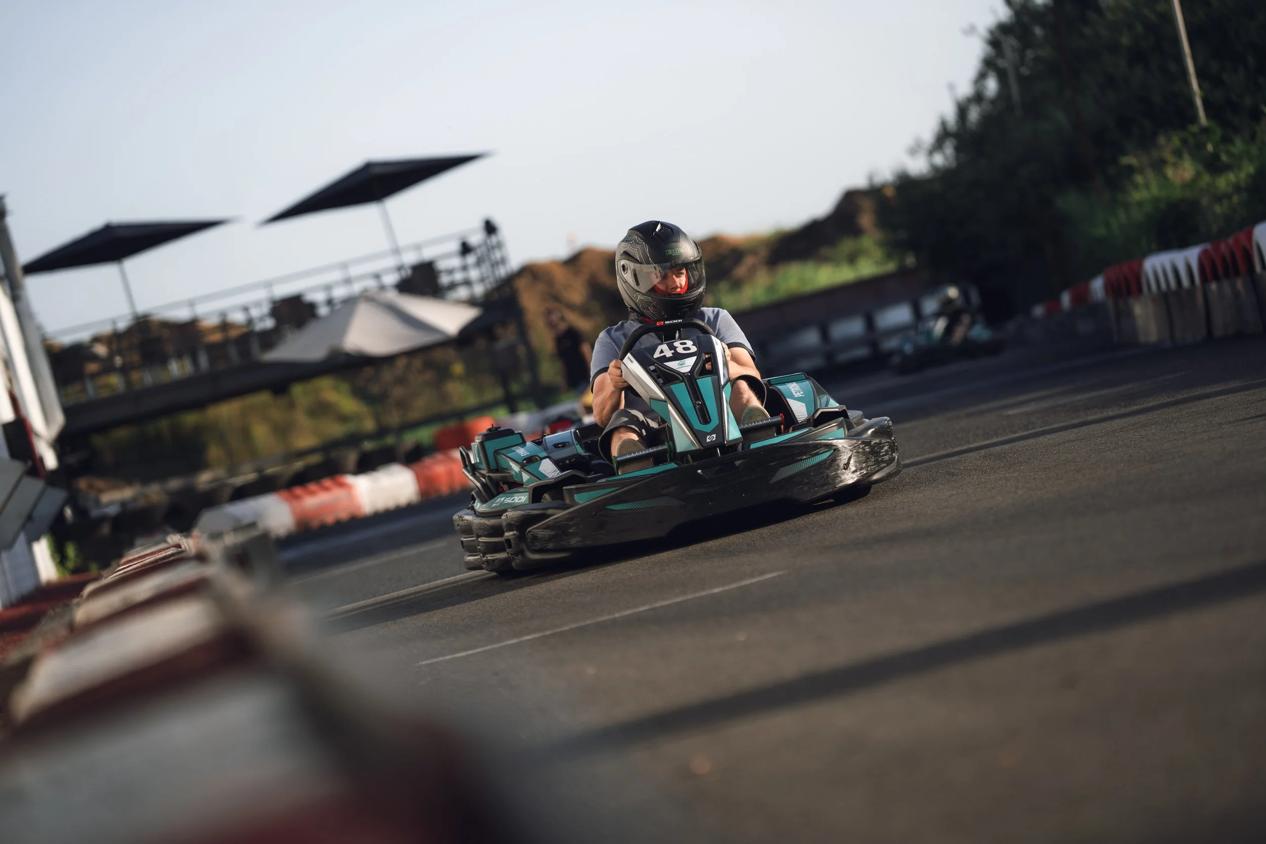 A person in a helmet and gray shirt riding a go-kart on a track, with safety barriers and trees in the background.
