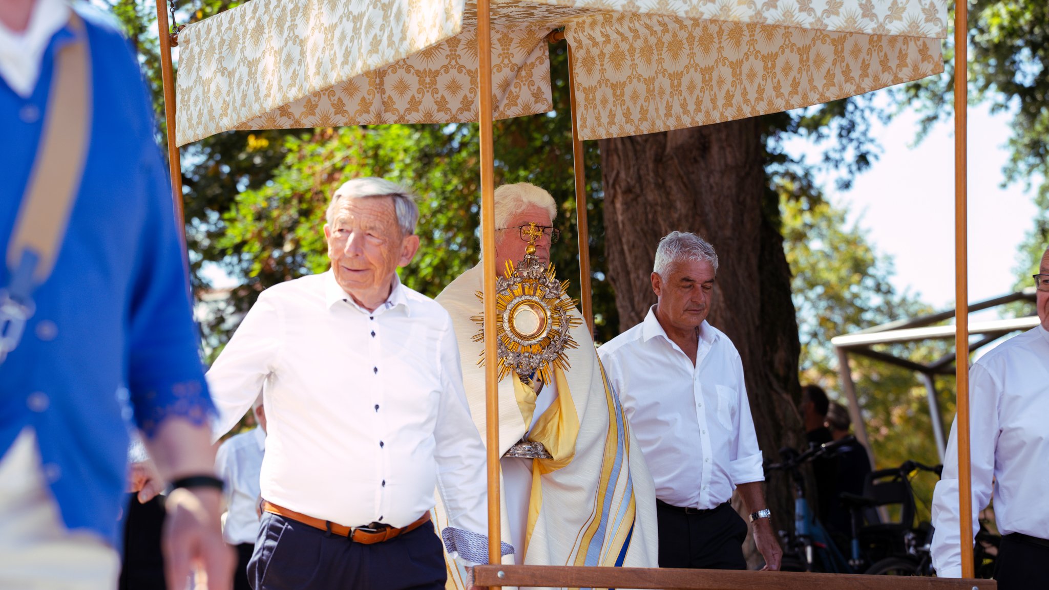 A group of elderly men standing under a canopy, participating in a religious or ceremonial event outdoors, with trees and blue sky in the background.