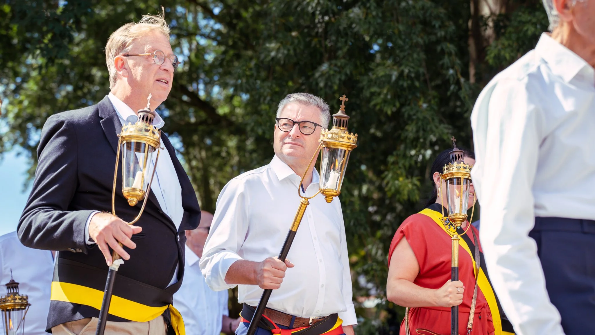 People participating in a solemn outdoor procession holding lanterns, with some wearing traditional ribbons and costumes, in a park with green trees.
