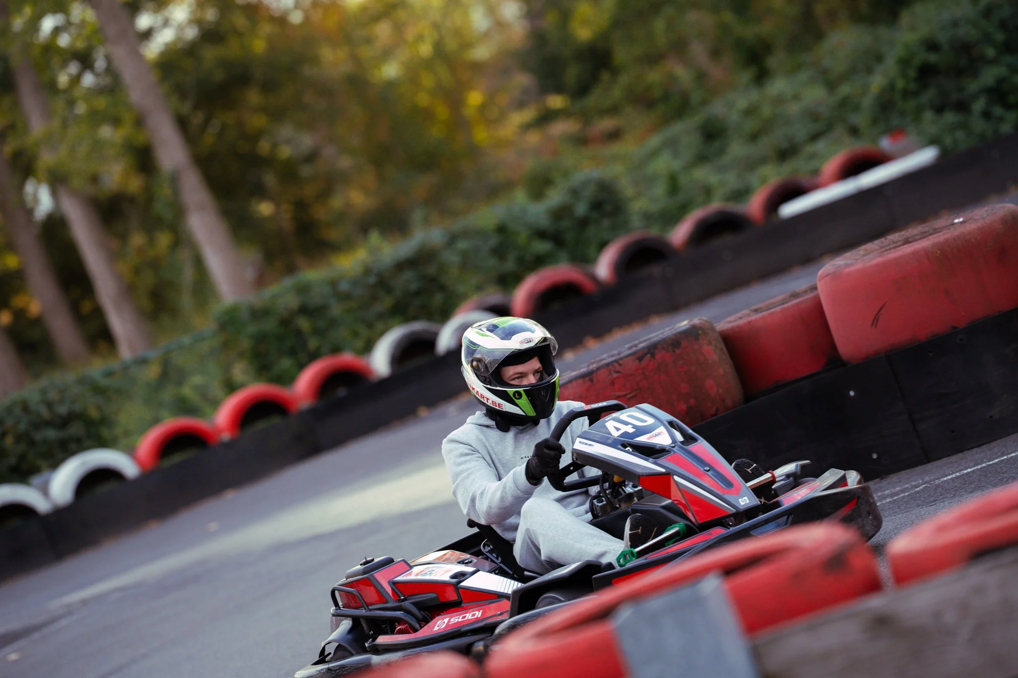 A person wearing a helmet and gray hoodie racing in a go-kart on a curving outdoor track with red and white barriers.