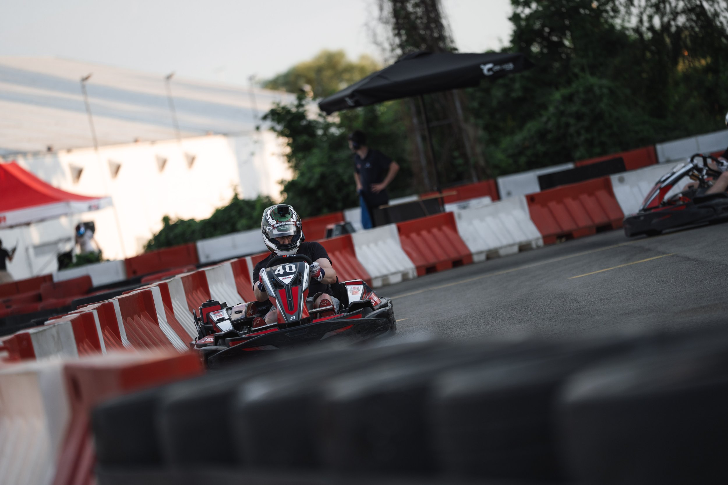 Person in a helmet driving a go-kart on an outdoor track with safety barriers, with another person standing in the background, trees, and a white building.