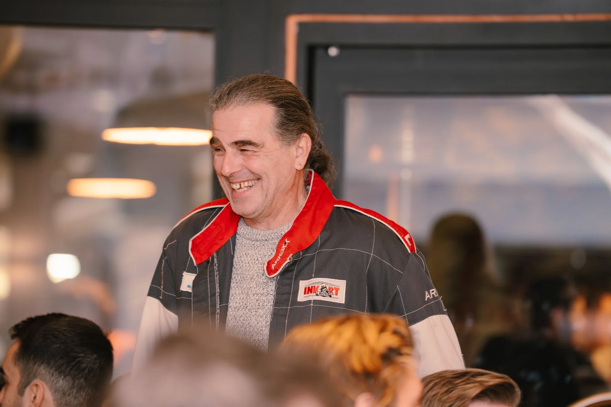 Man in racing jacket smiling in a casual indoor setting, with people seated in front of him.