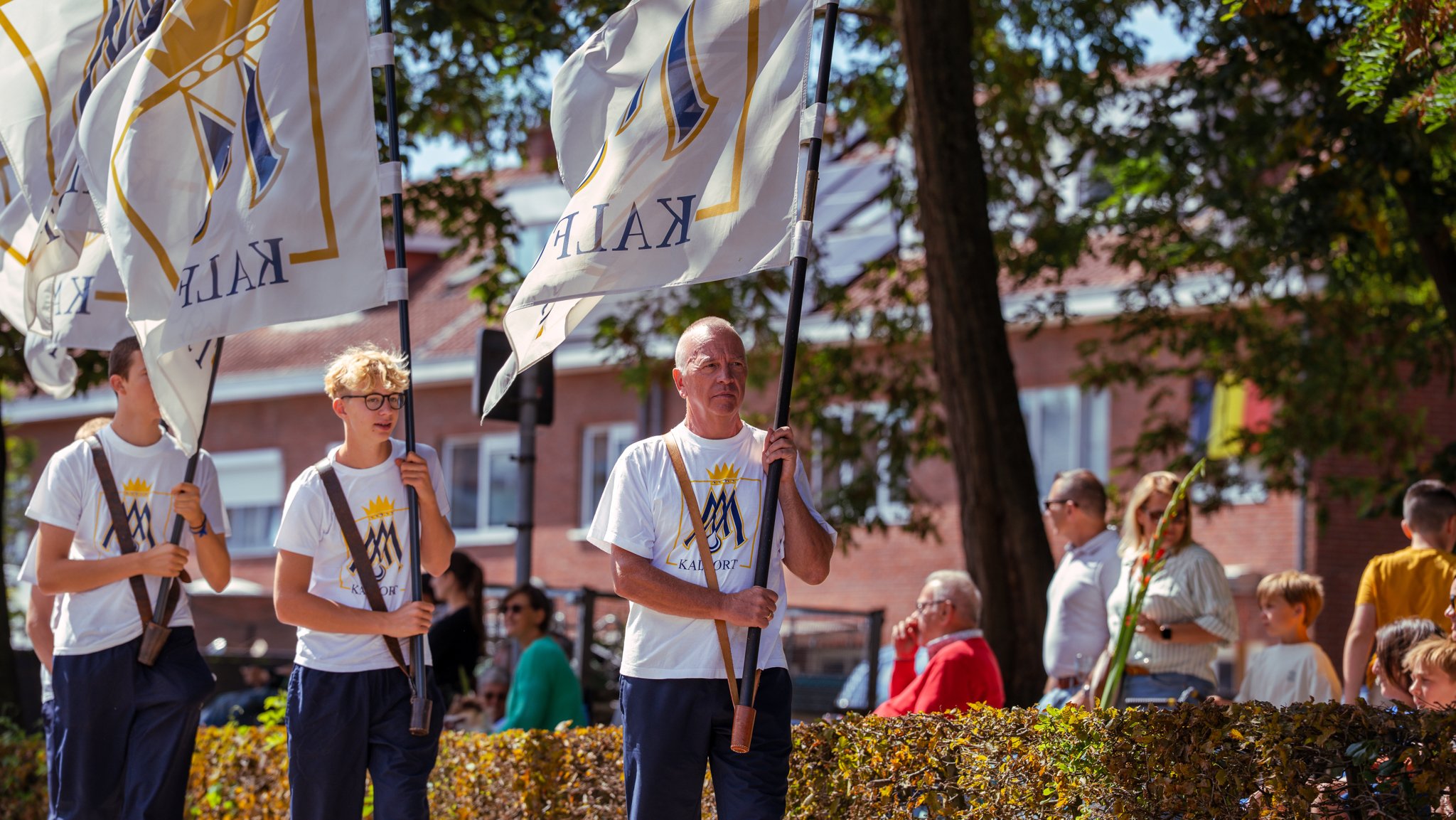 People holding flags during an outdoor event, with a man and two boys in white T-shirts with a logo, standing behind a hedge. Others are in the background, standing near trees and brick buildings.
