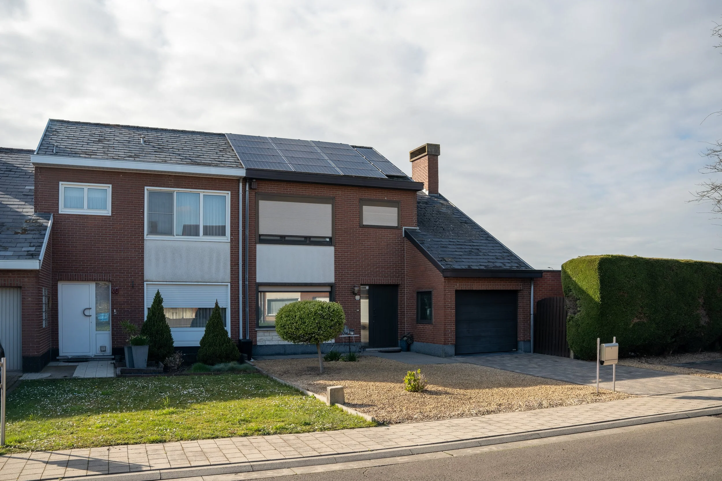 A two-story brick house with solar panels on the roof, surrounded by a small front yard with grass, a tree, and bushes. There is a driveway with a garage door on the right and a pathway leading to the front door on the left.